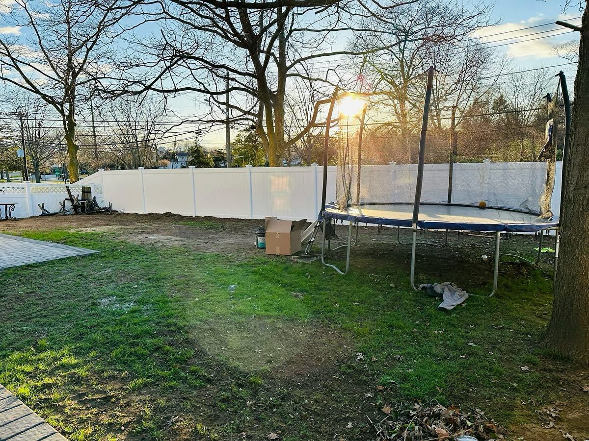 Backyard with trampoline, white fence, and trees. Green grass and sunlight.