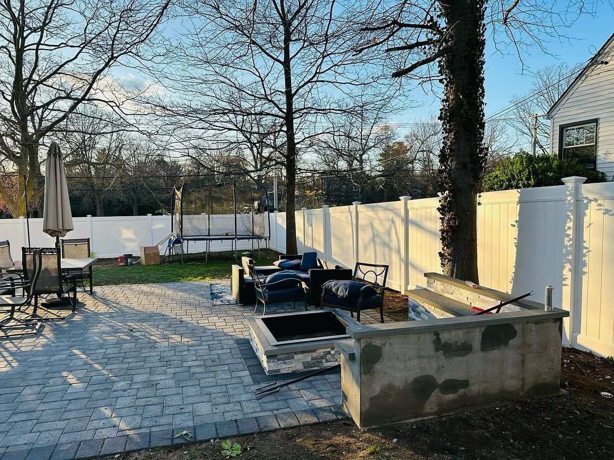 Backyard patio with fire pit, seating, trampoline, and white fence.