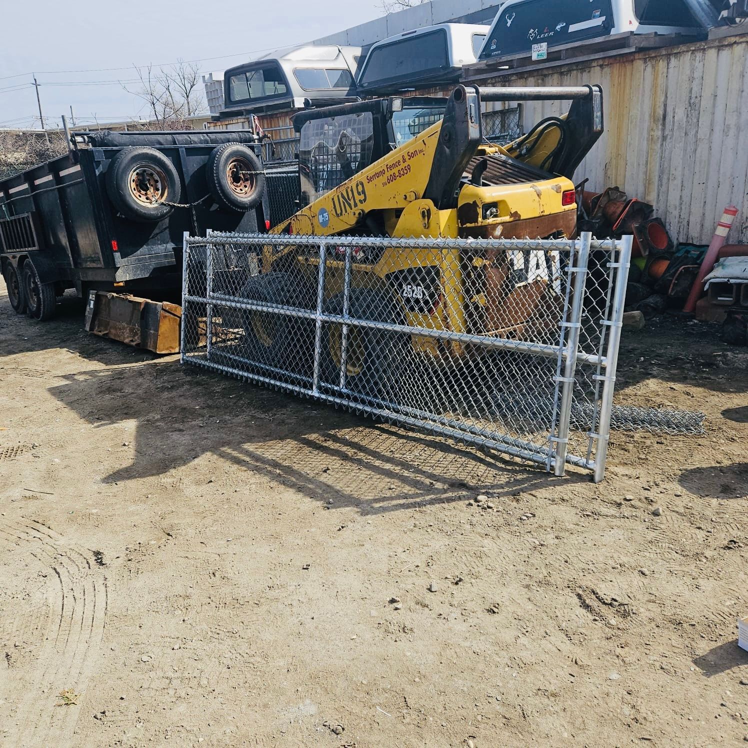 Chain link gate on the ground, with a trailer, skid steer, and other machinery in a storage yard.