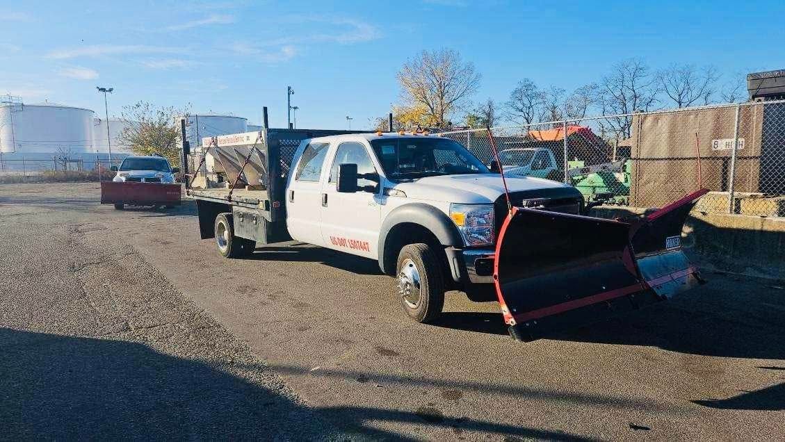 White truck with snow plow on a gravel lot under a blue sky, another truck in background.