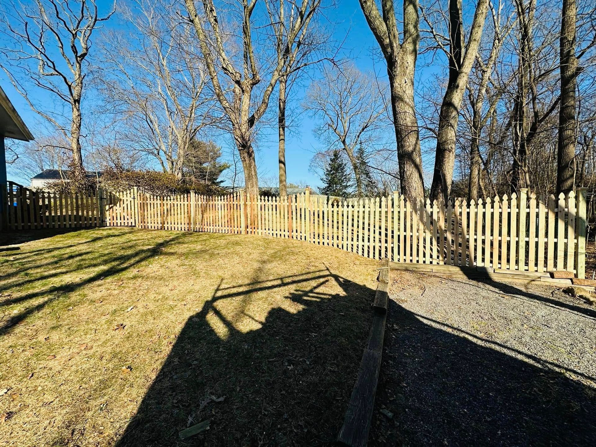 Backyard with picket fence, trees, and gravel path on a sunny day.