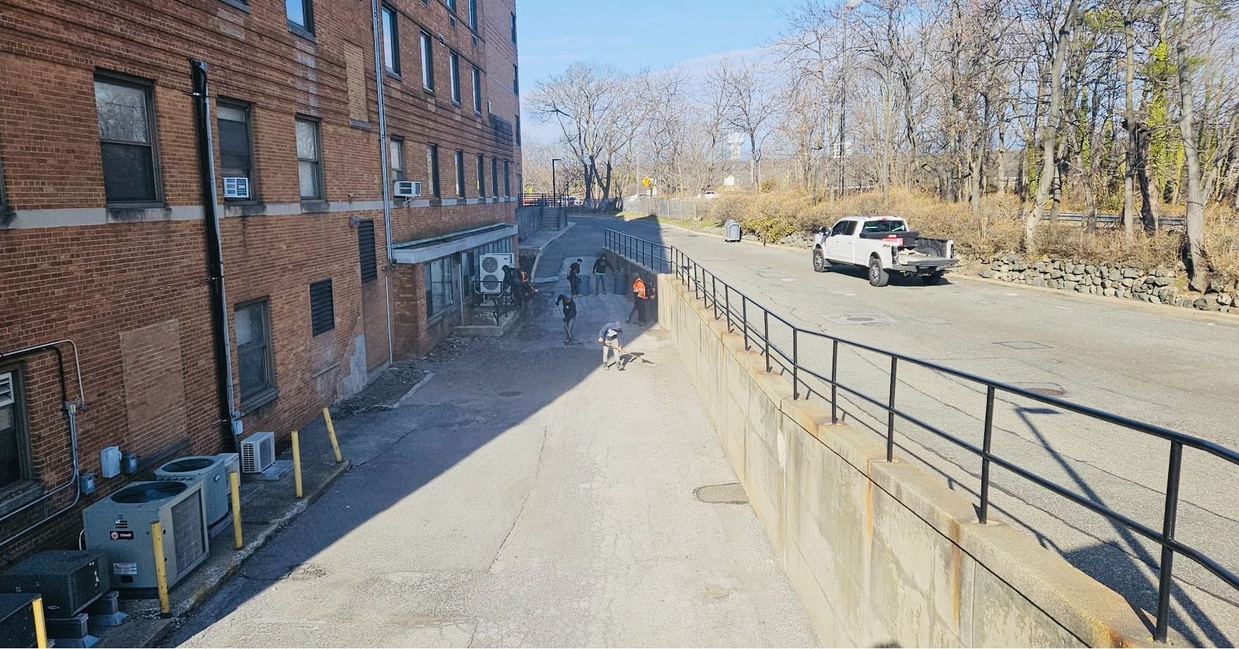 Back alley with a building, people, and a truck. Bright sun, concrete, black railing, and trees.
