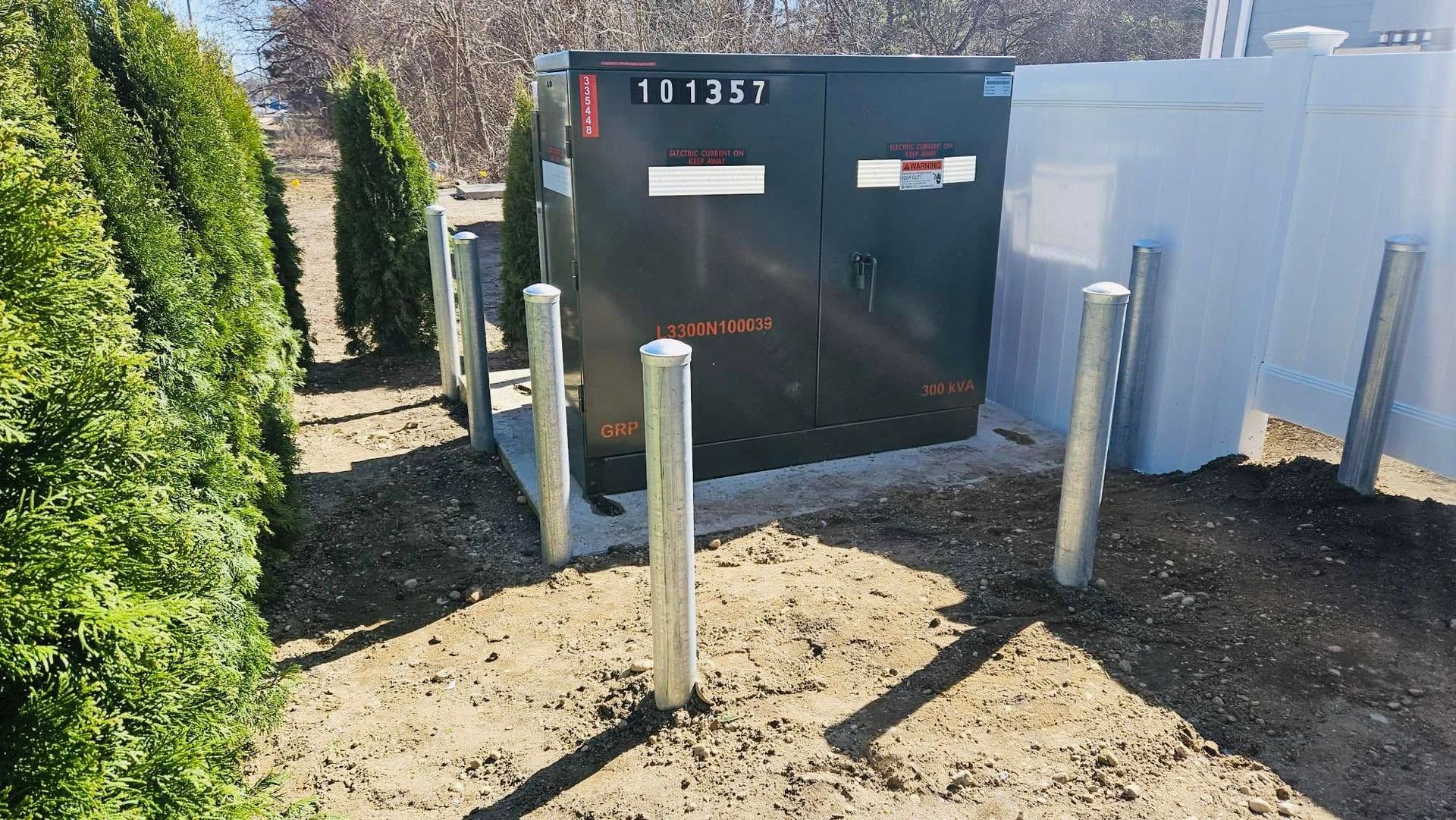 Black electrical box with metal bollards in front, near green bushes and a white fence.