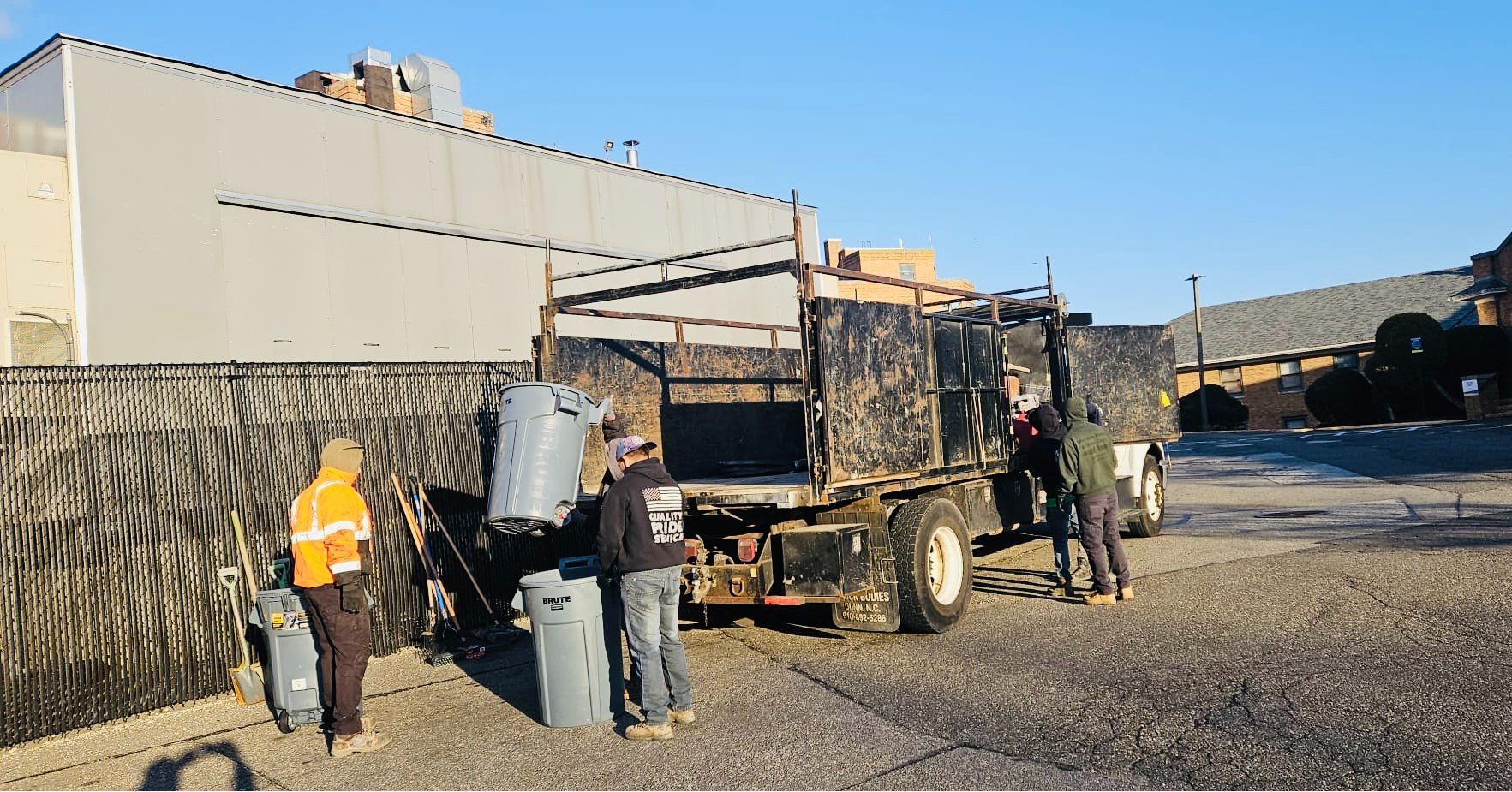 People loading gray trash cans into a truck in a paved lot, beside a tall building and fence.