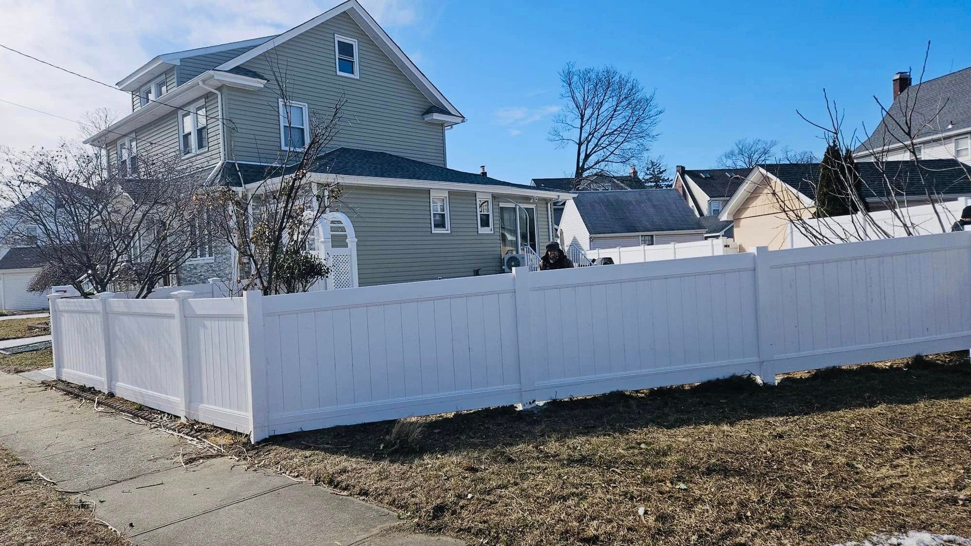 White fence surrounds a two-story house with siding. The sky is blue and the ground has patches of snow.