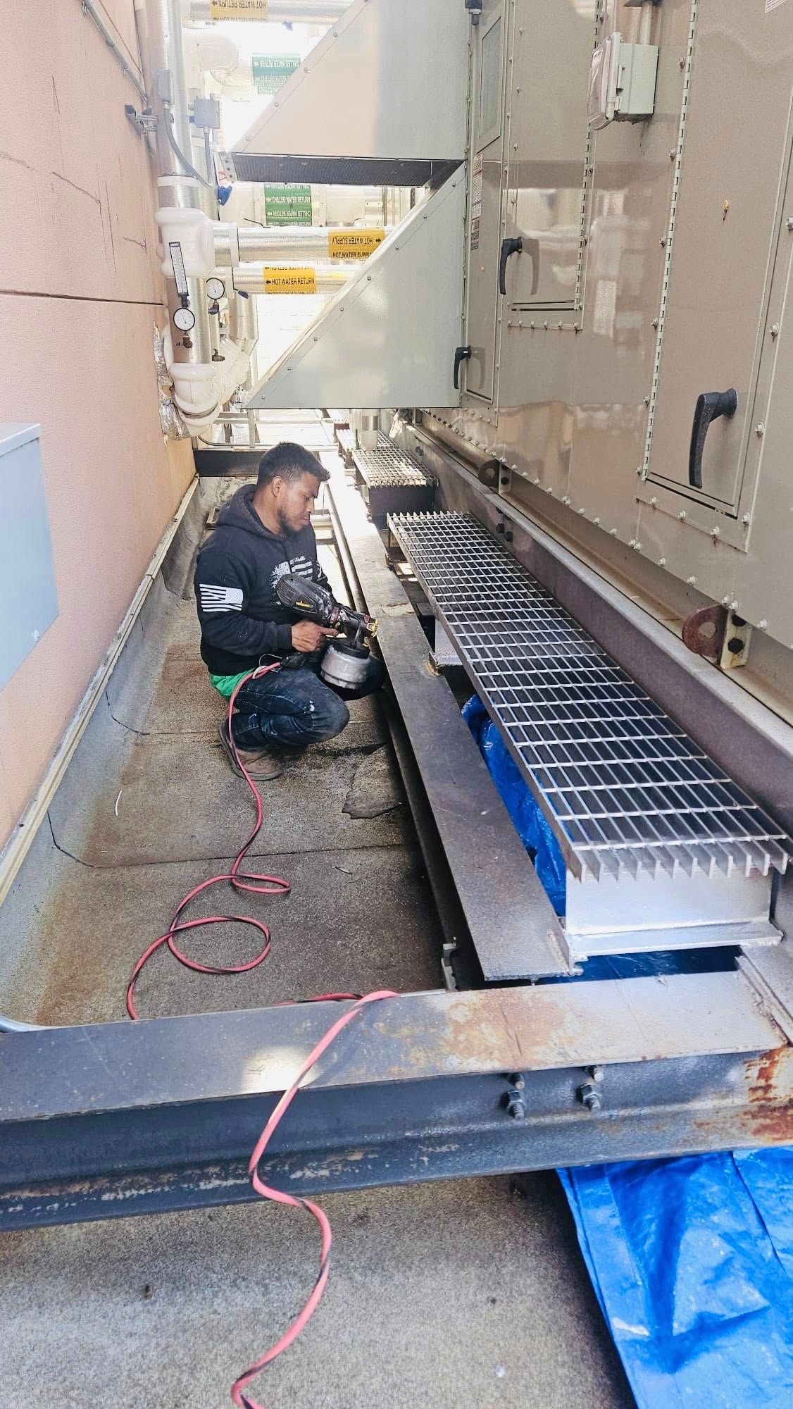 Person working on machinery. Metal grating and blue tarp visible. Outdoors setting.