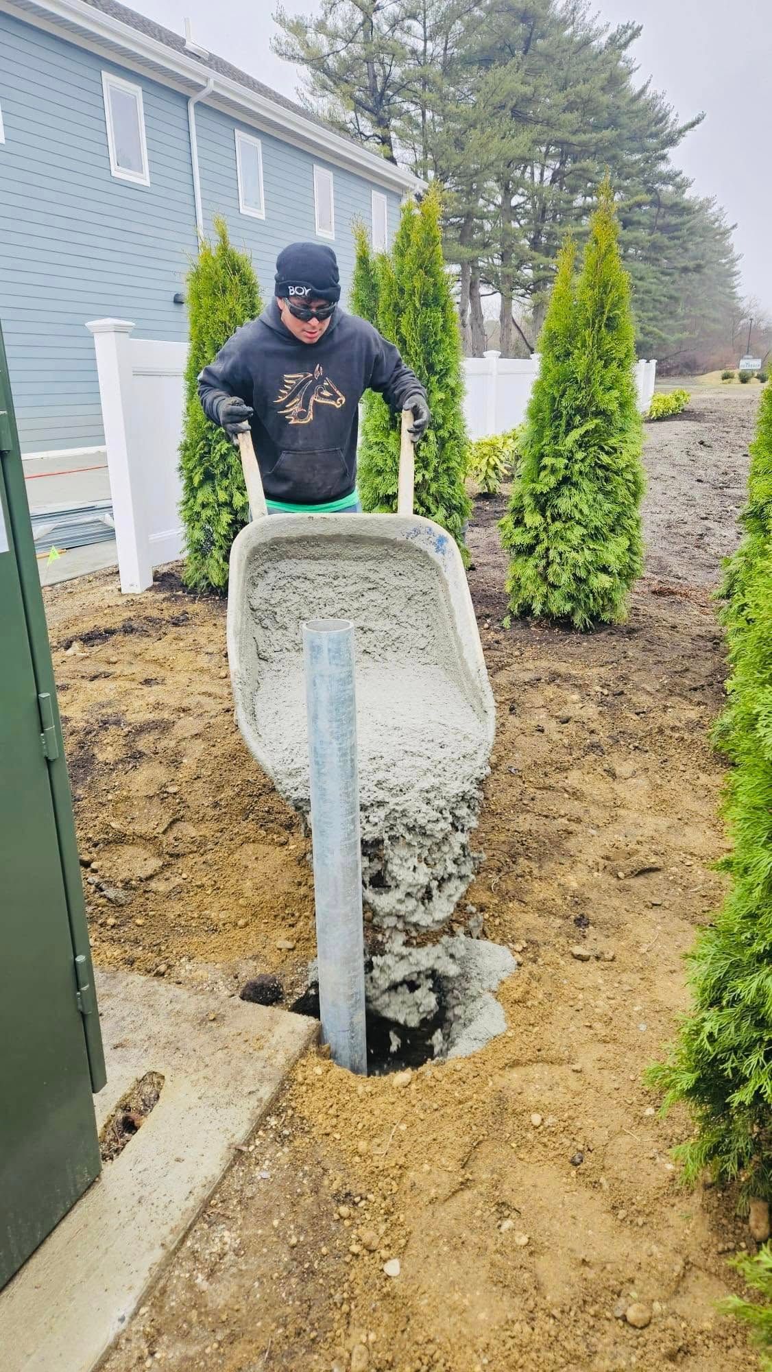 Man pouring concrete from wheelbarrow into a hole around a post in a yard.