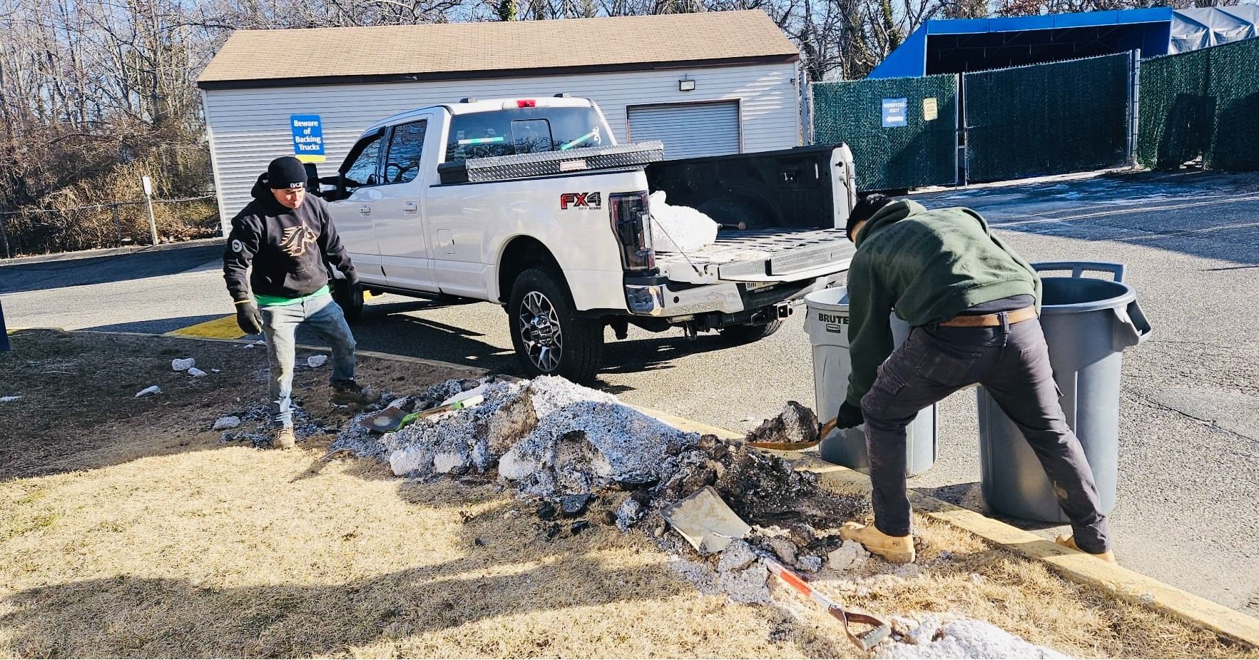 Two people cleaning up debris next to a pickup truck and two trash cans outside a building.