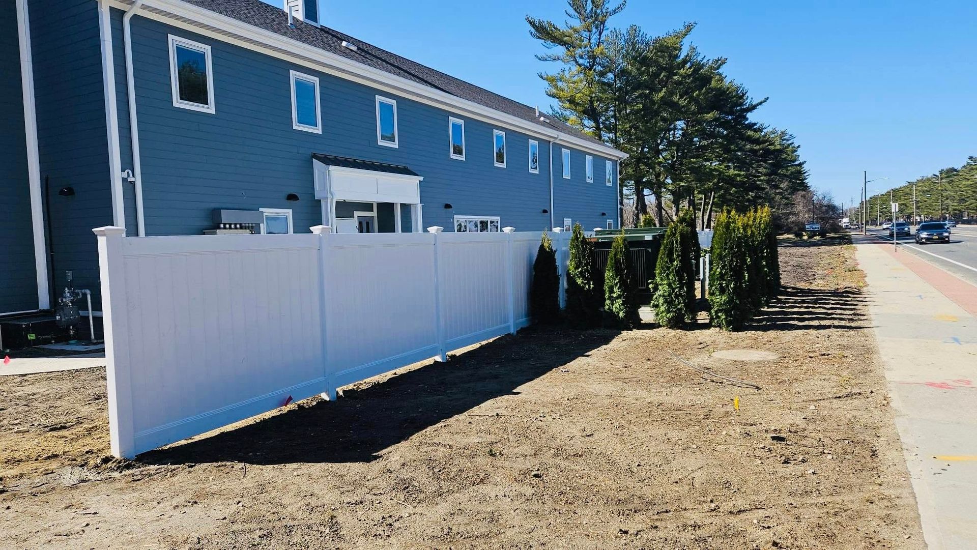 White fence in front of a blue building with several windows. Brown dirt and a sidewalk are in front.