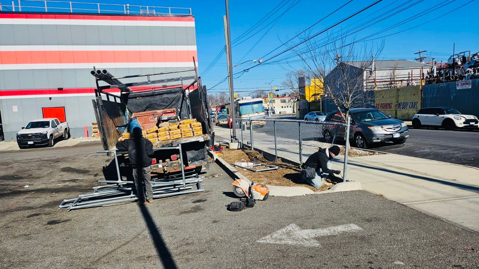 Truck unloading debris, two people working near a fence. Gray car and white building in the background. Clear sky.