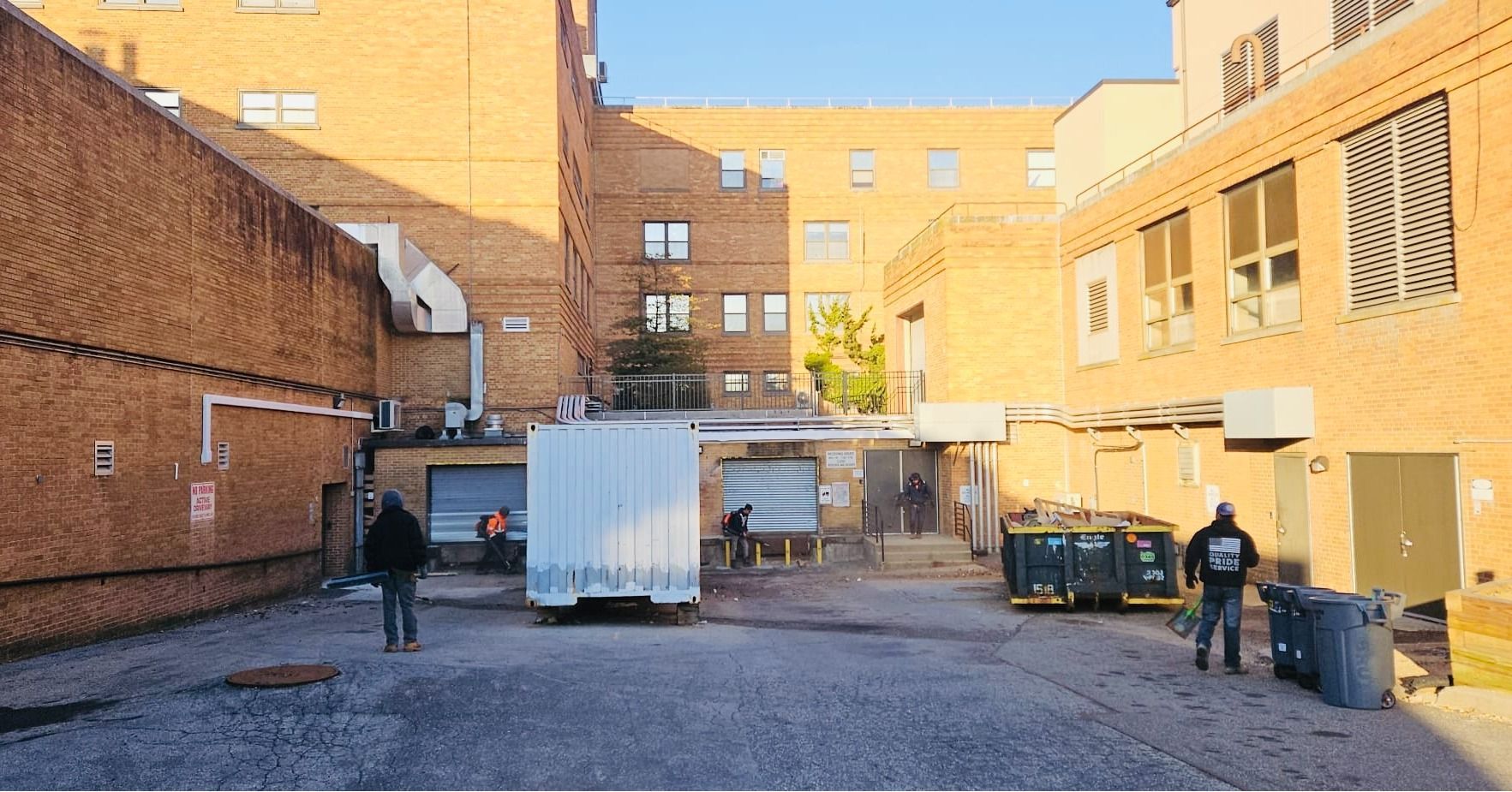 Brick buildings and a loading dock with people near a white container, blue bins, and a closed door.