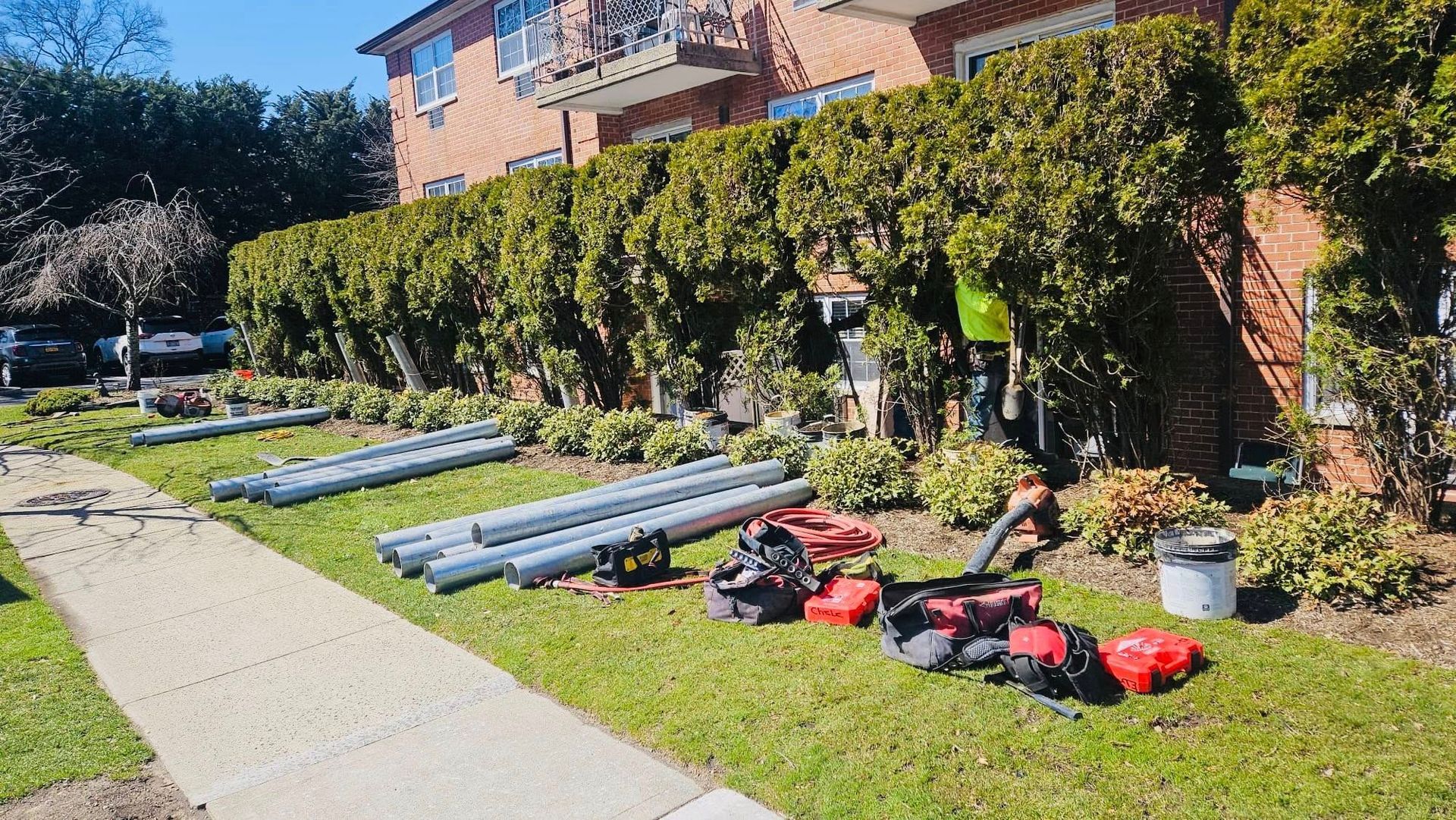Lawn with long, gray pipes, tools, and a worker trimming hedges next to a brick building and sidewalk.