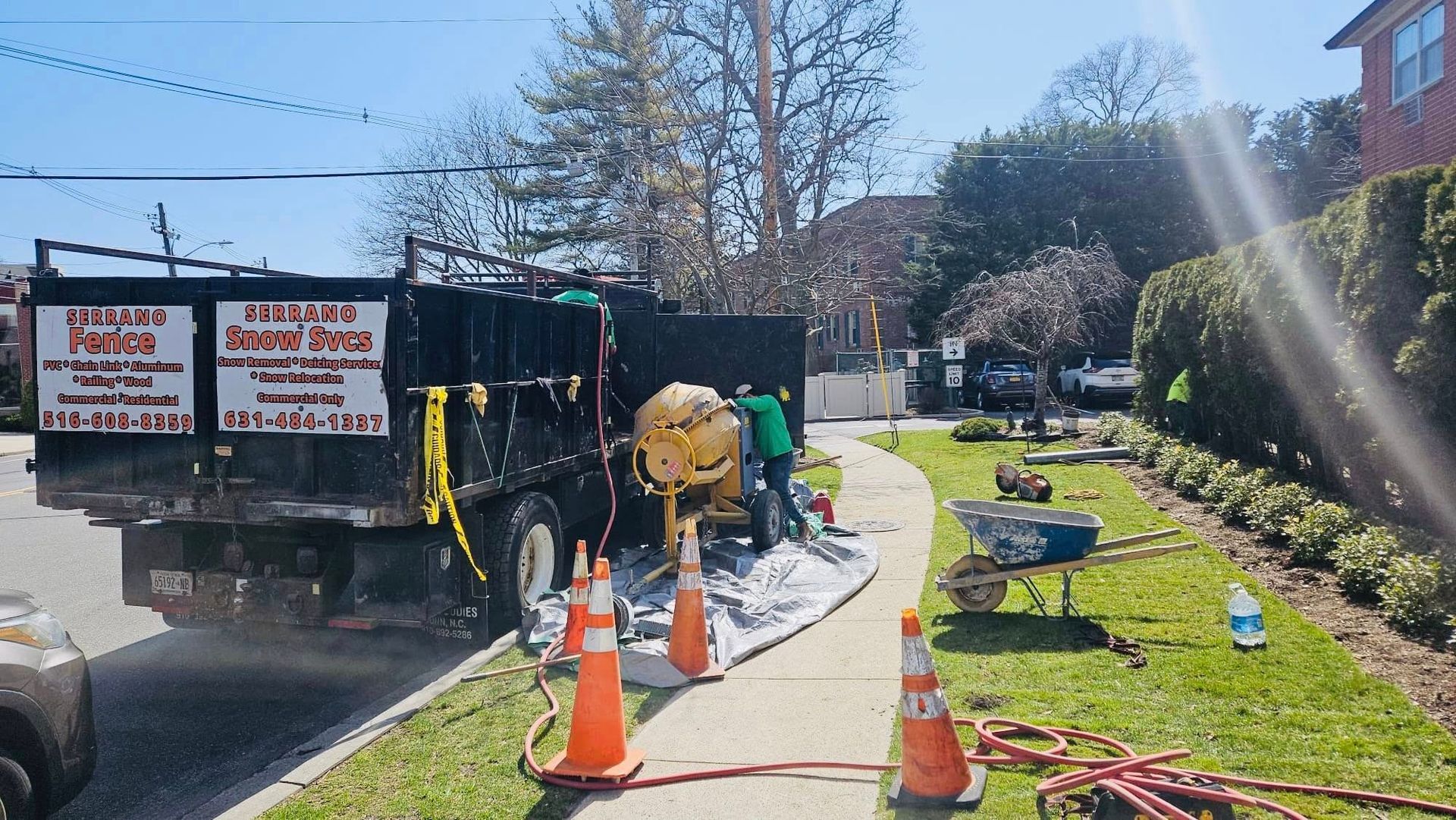 Construction workers mixing cement on a sidewalk next to a truck. Orange cones and hoses are present.