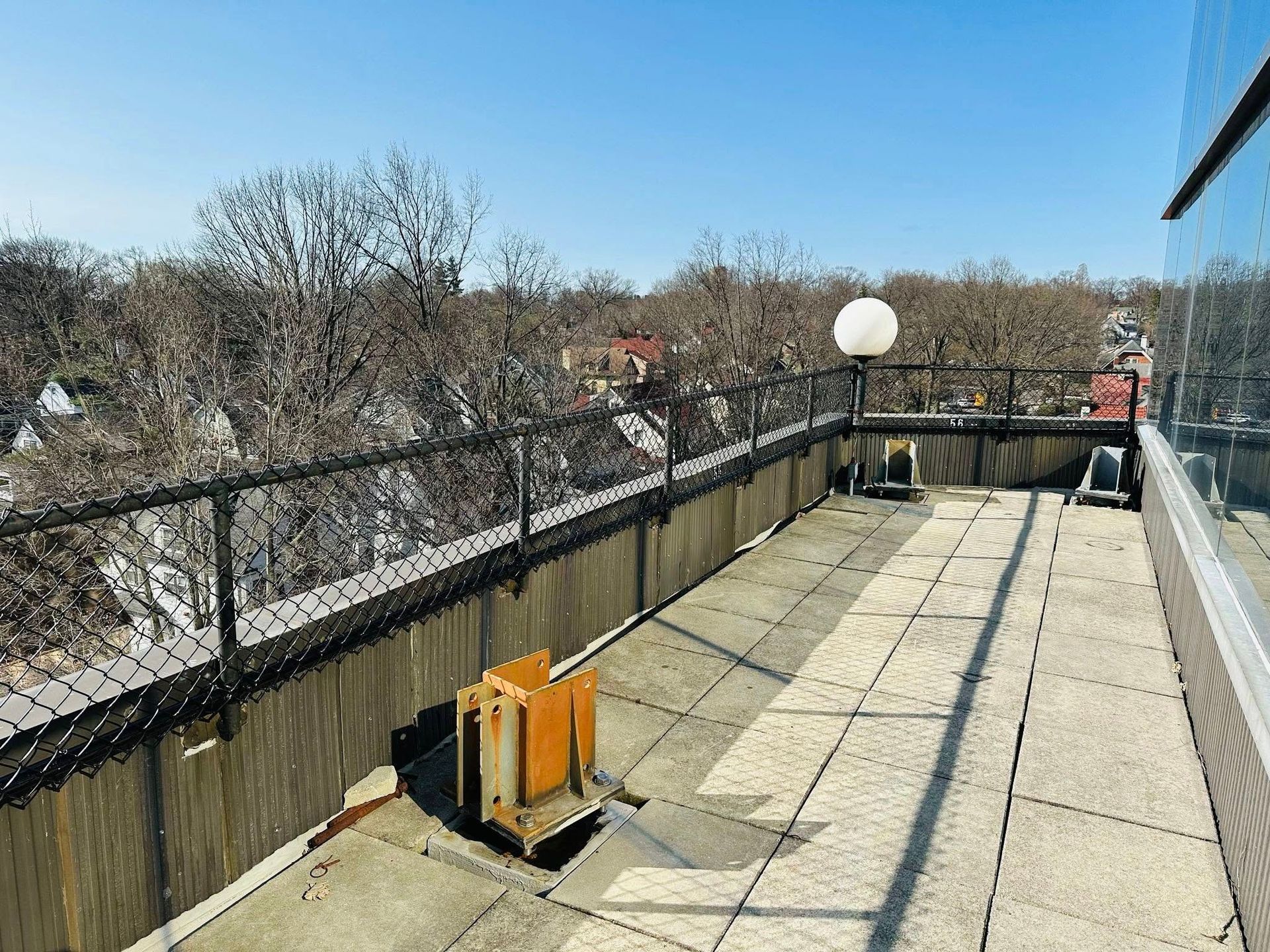 Rooftop view with chain link fence, concrete, and surrounding trees under a clear blue sky.
