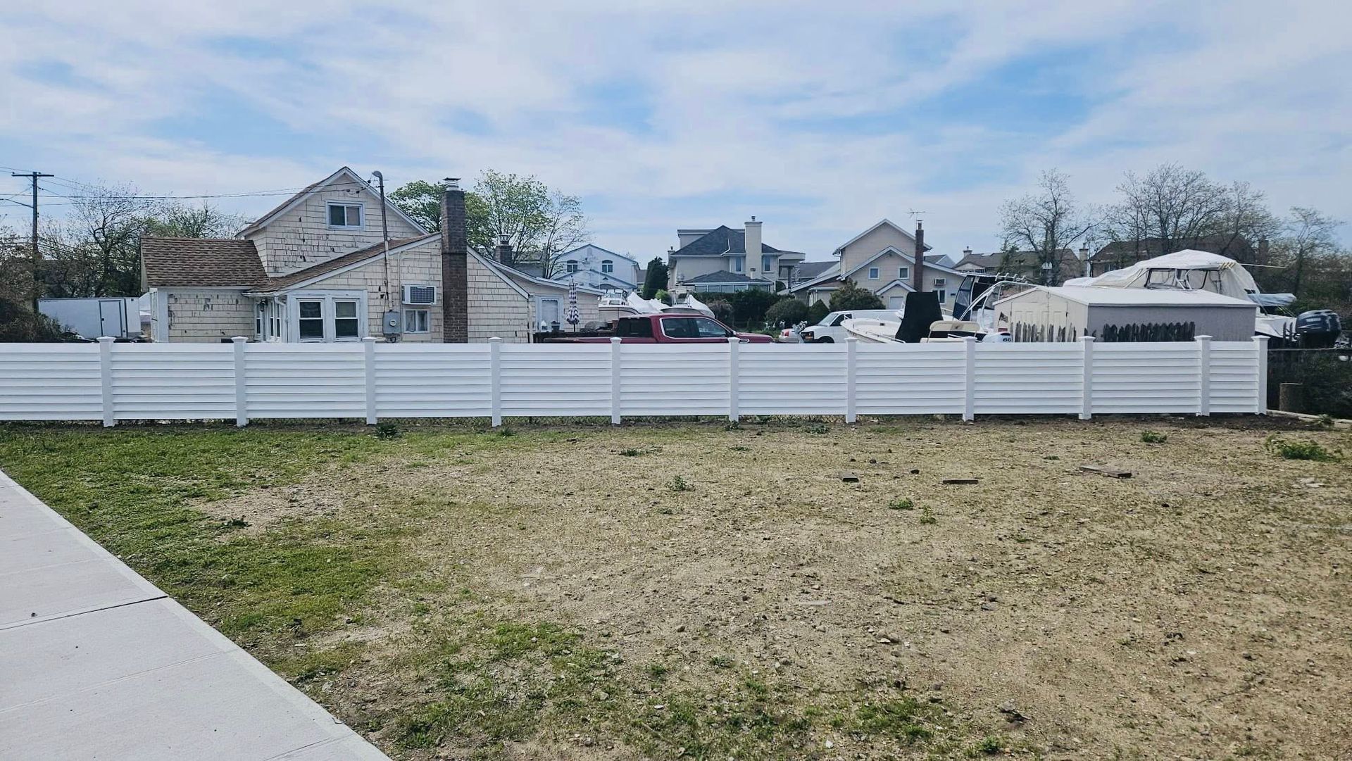 White fence in front of a vacant lot; houses and a blue sky in the background.