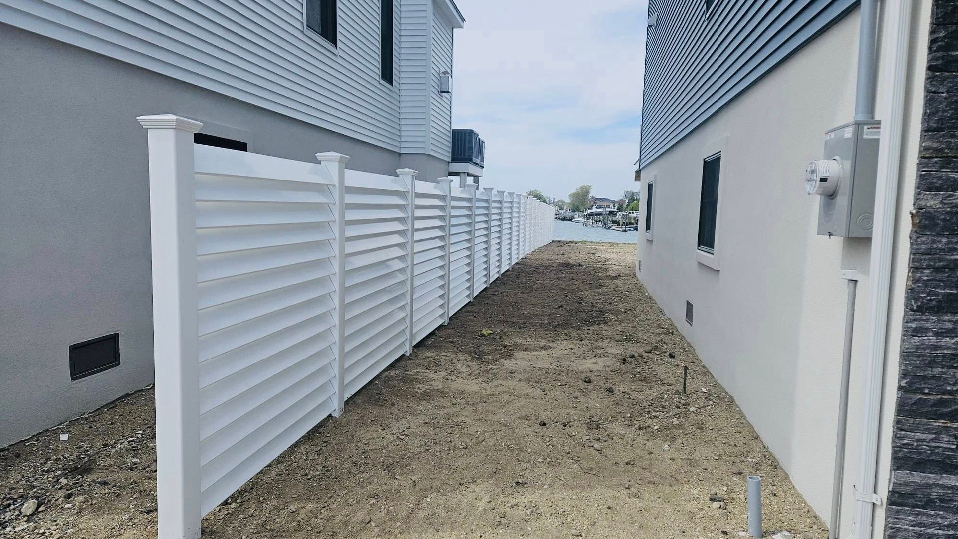 White privacy fence between two houses, on a dirt path with a view of water and sky.