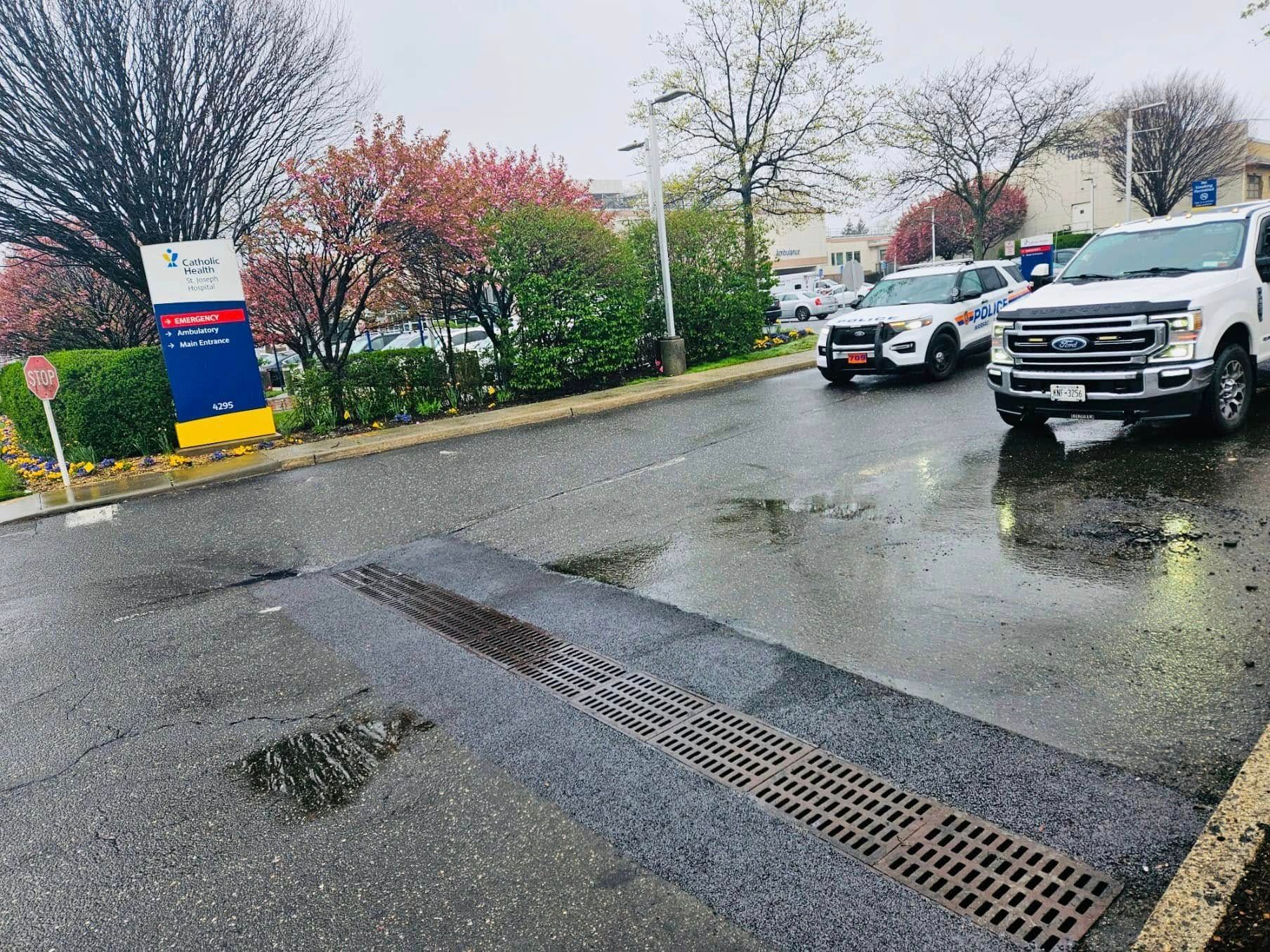 Entrance to a hospital with two white trucks. Sign indicates emergency services. Road has drainage grates. Cloudy day.