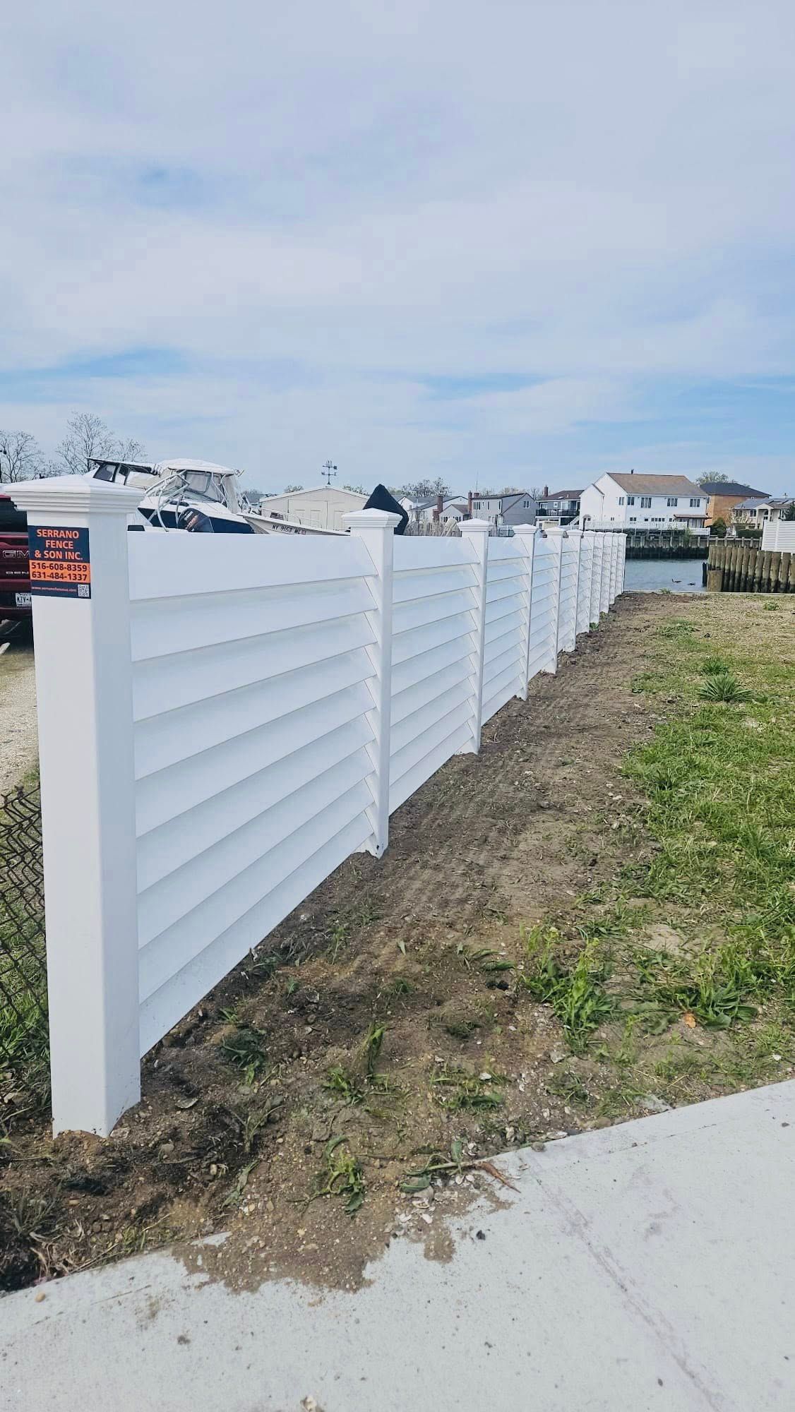 White vinyl fence along dirt and grass, with buildings in the background under a blue sky.