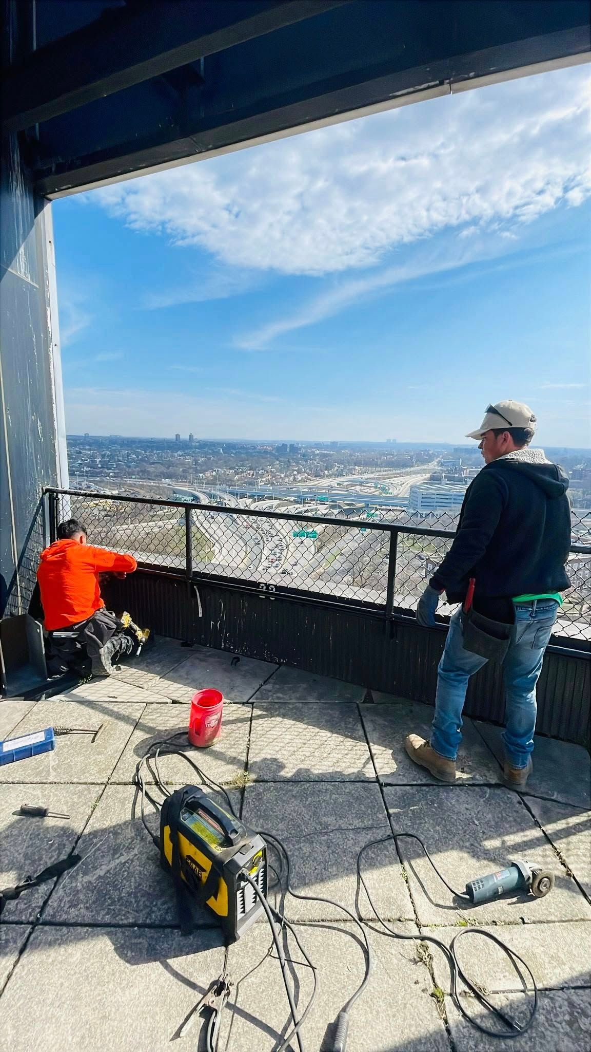 Two construction workers on a rooftop patio overlooking a cityscape under a blue sky; tools scattered around.