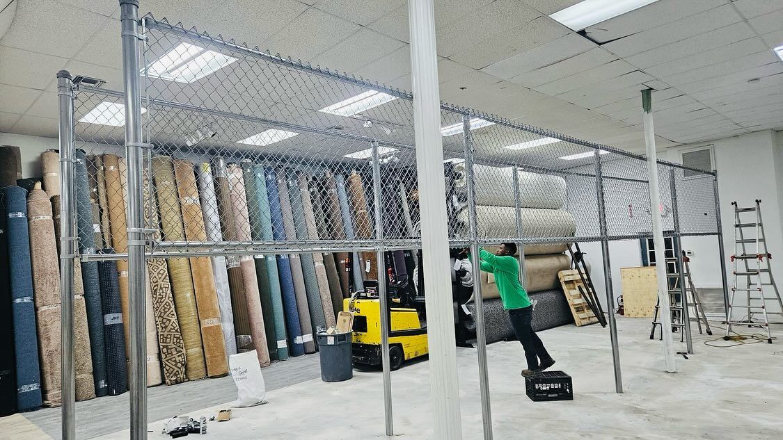 Warehouse with rows of carpet rolls, worker on a crate near a yellow forklift, metal shelving.