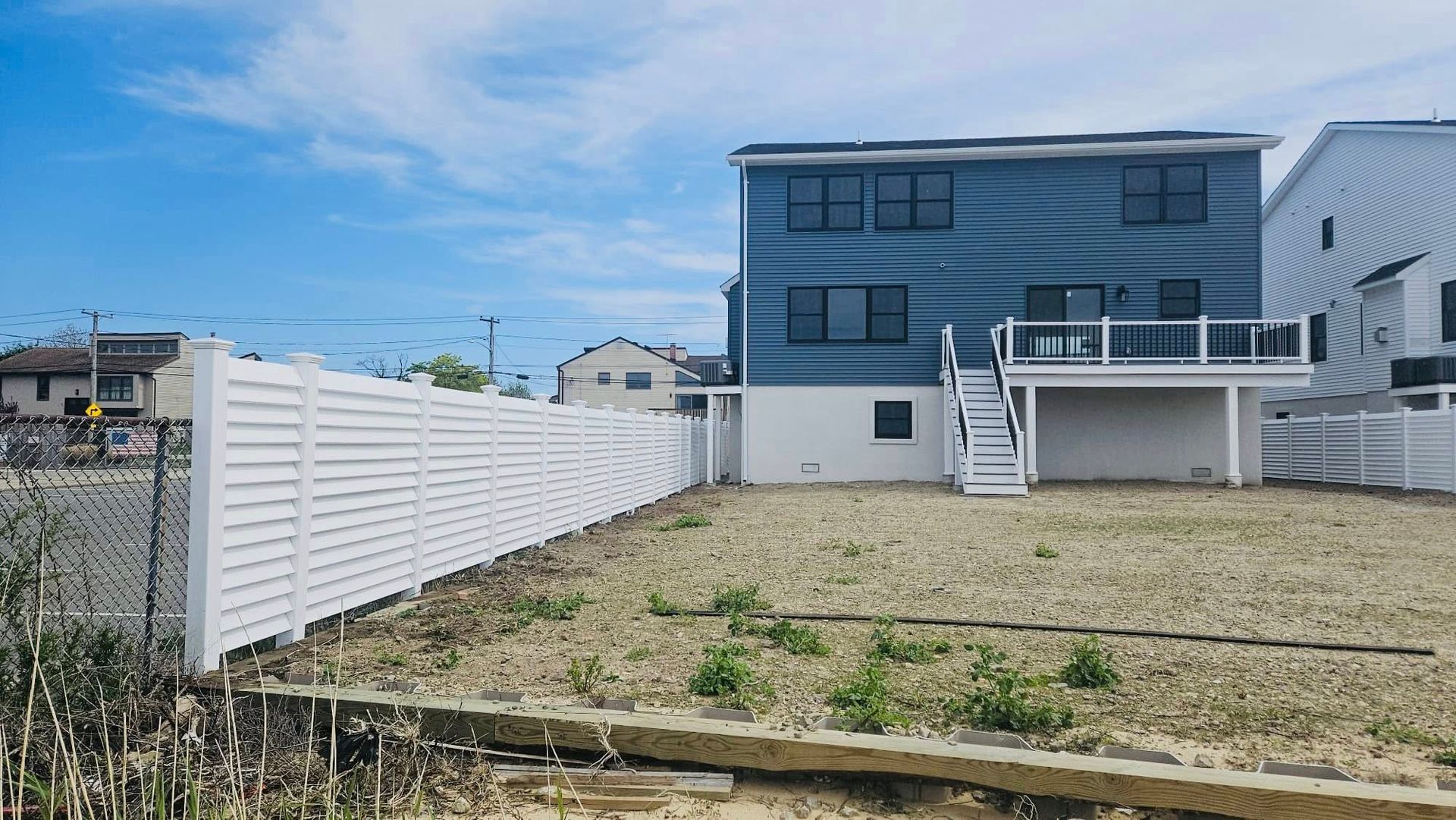Two-story blue house with white deck and fence, on a brown dirt lot under a blue sky.
