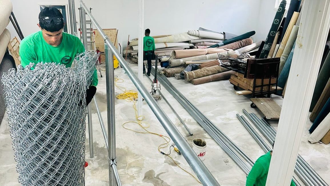 Man in green shirt rolls out chain-link fence in a room with construction materials.