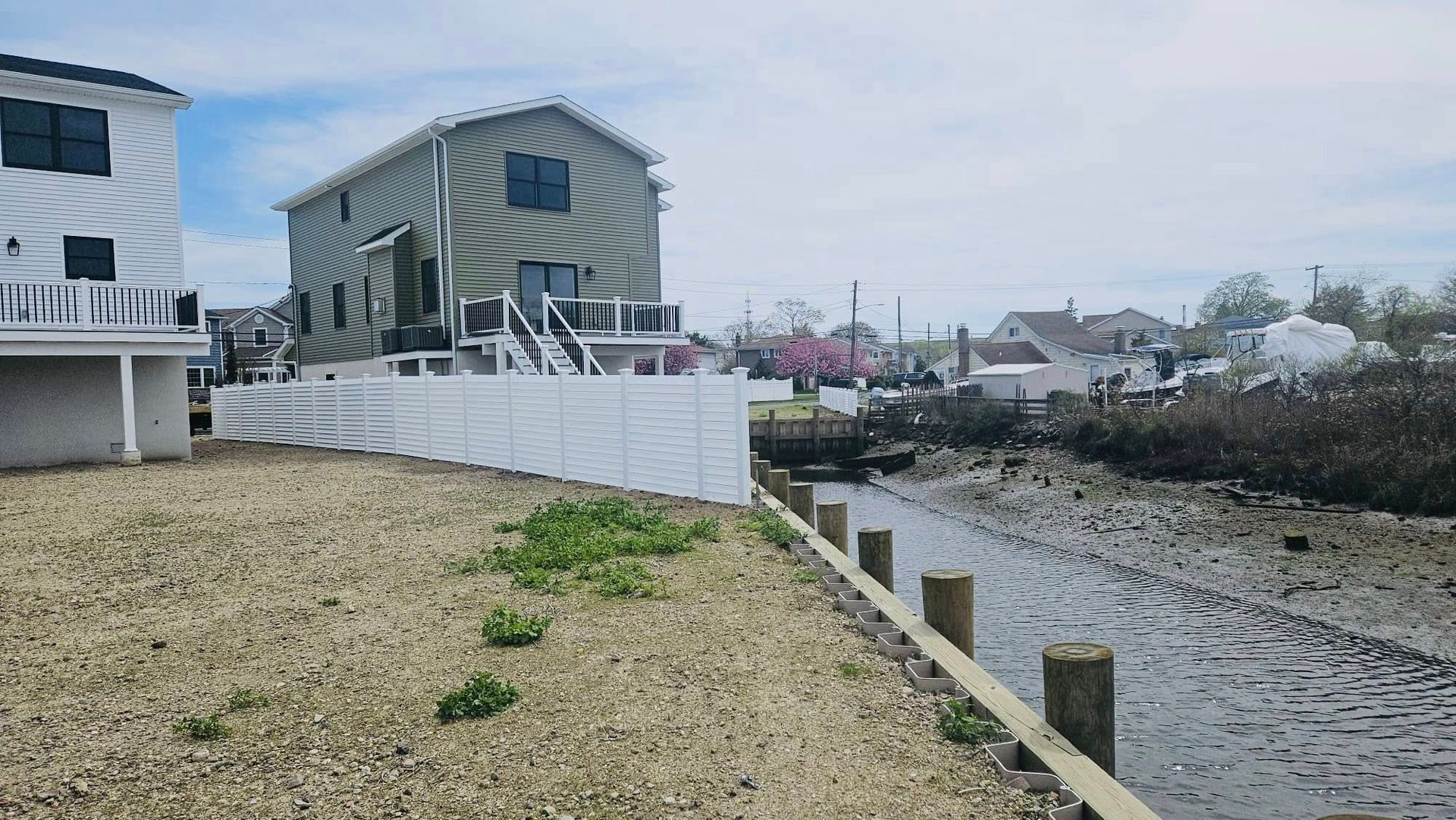 A coastal scene: houses on a canal with muddy banks, a white fence, and a cloudy sky.