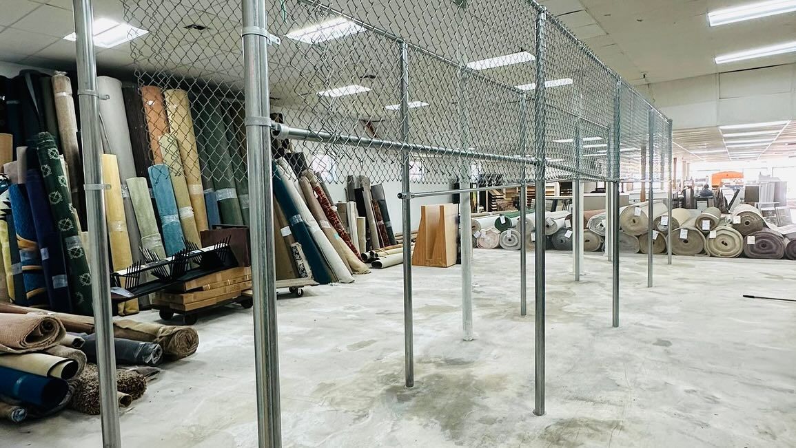 Warehouse with fabric rolls along the left wall, enclosed by a chain-link fence, and metal posts.