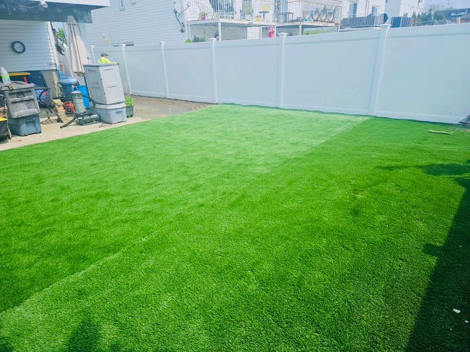 Lush green artificial turf in a fenced backyard, bright white fence in the background.