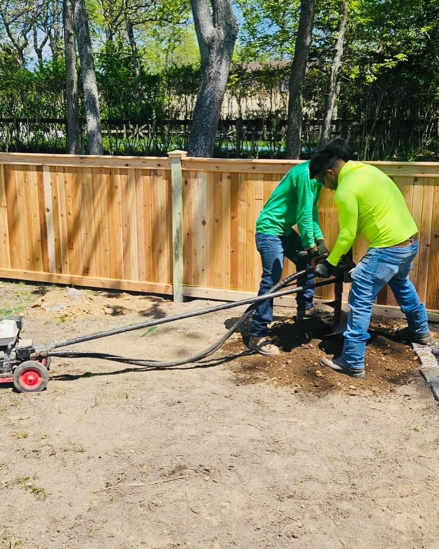 Two workers in green and yellow shirts using a motorized auger to drill a hole near a wooden fence.