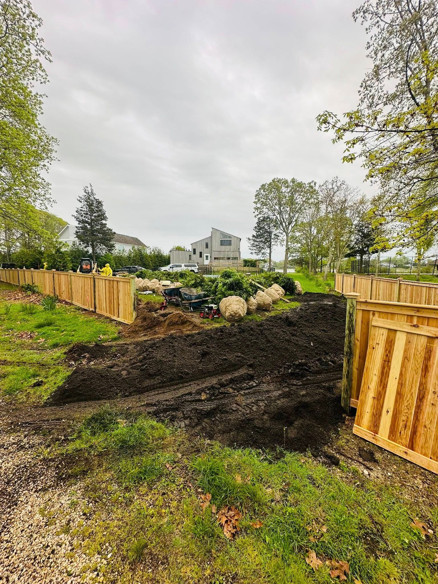 A wooden fence surrounds a garden with a pile of dark soil and a house in the background under a cloudy sky.