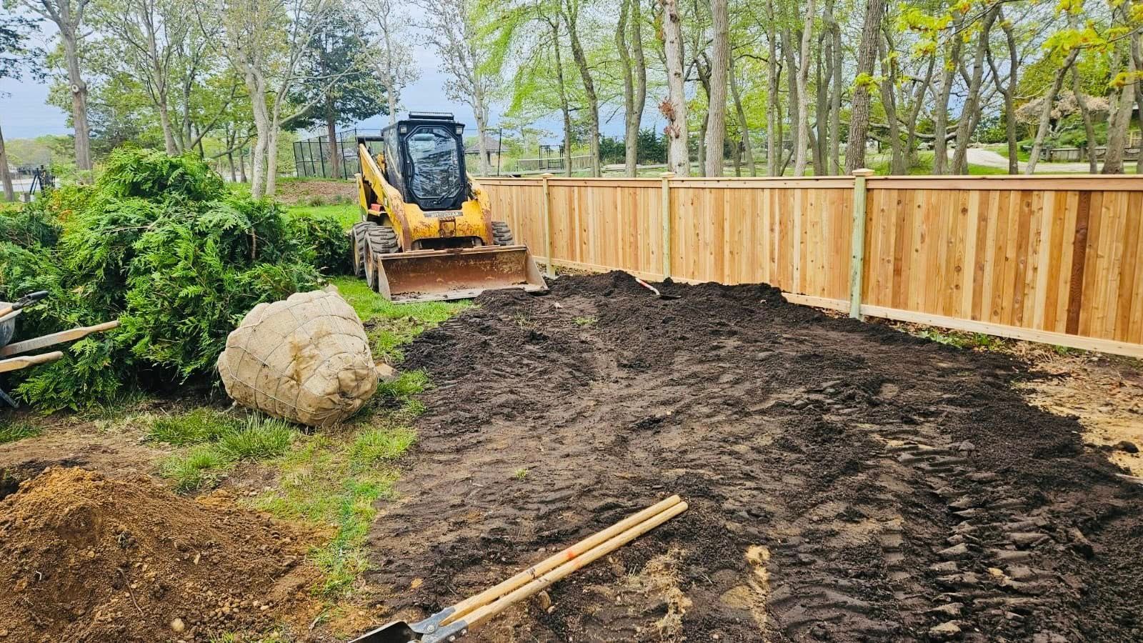 Skid steer moves dirt near a wooden fence and trees. A large pile of dirt is in the foreground.
