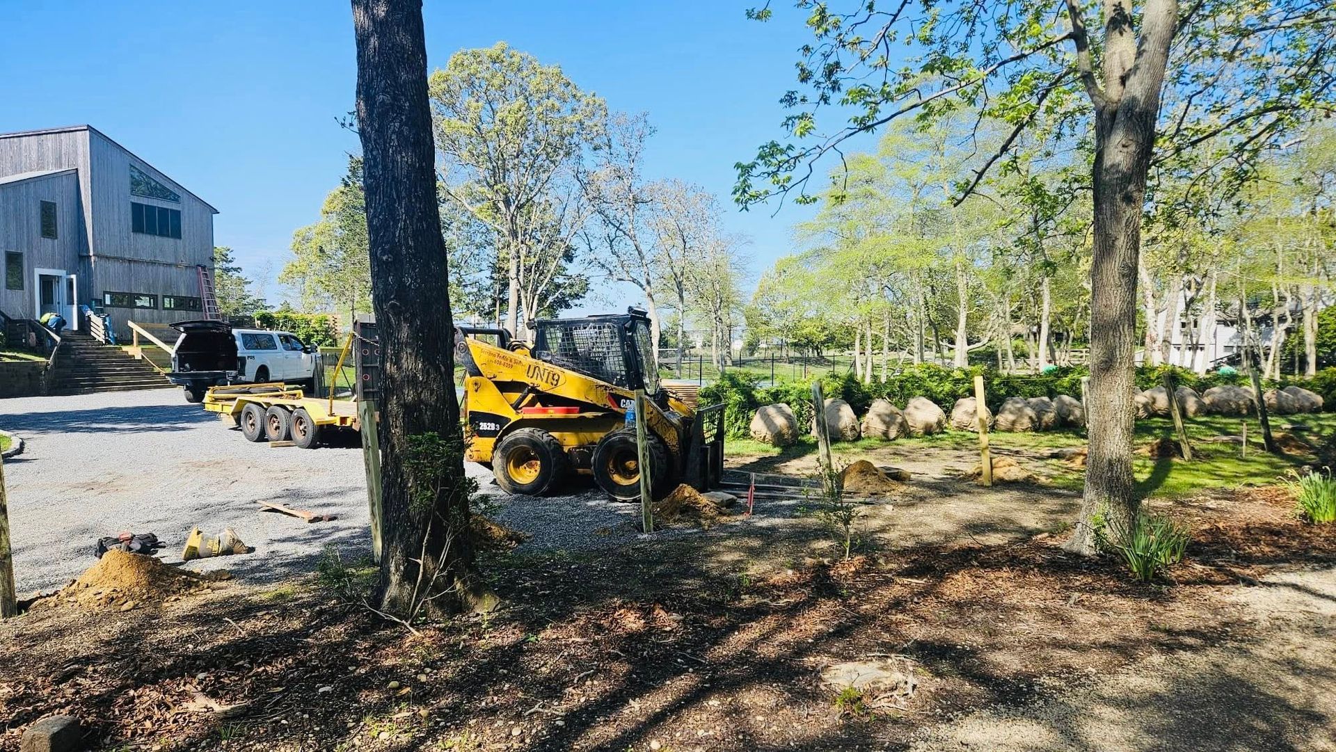 Yellow skid steer on a gravel driveway in front of a modern house. Trees surround the area.