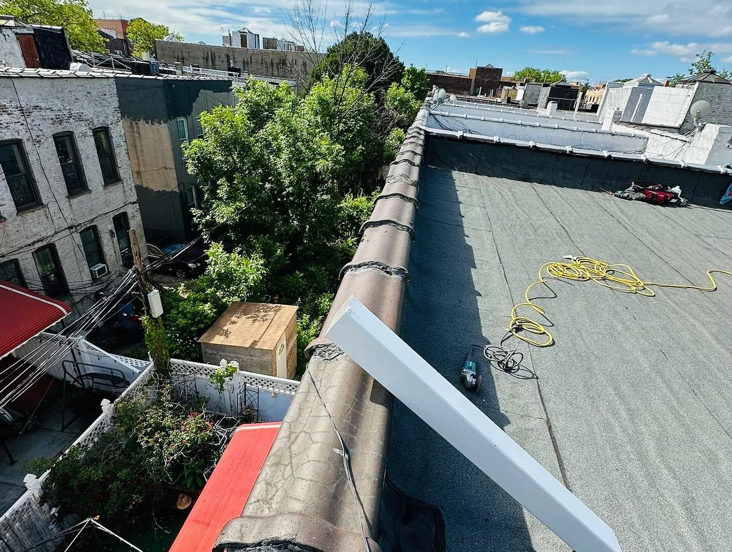 Rooftop view of gray rooftops, green trees, and blue sky. A white panel is on a roof edge.