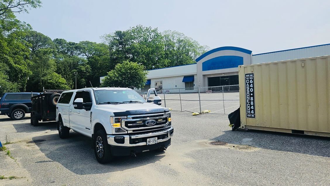 White pickup truck parked in front of a building with a blue awning and a shipping container; sunny day.