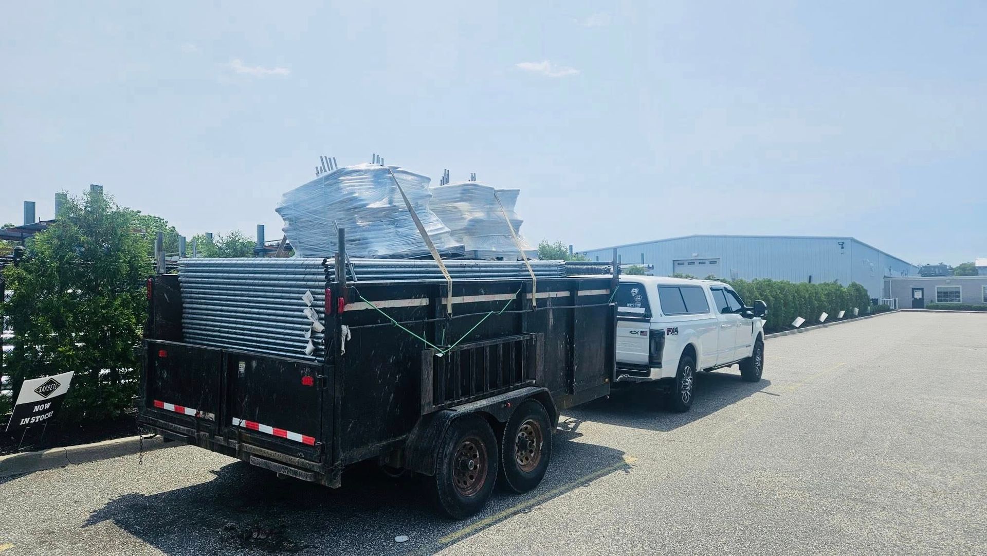 White truck towing a trailer loaded with metal piping and stacked sheet metal on a gravel lot near a factory.