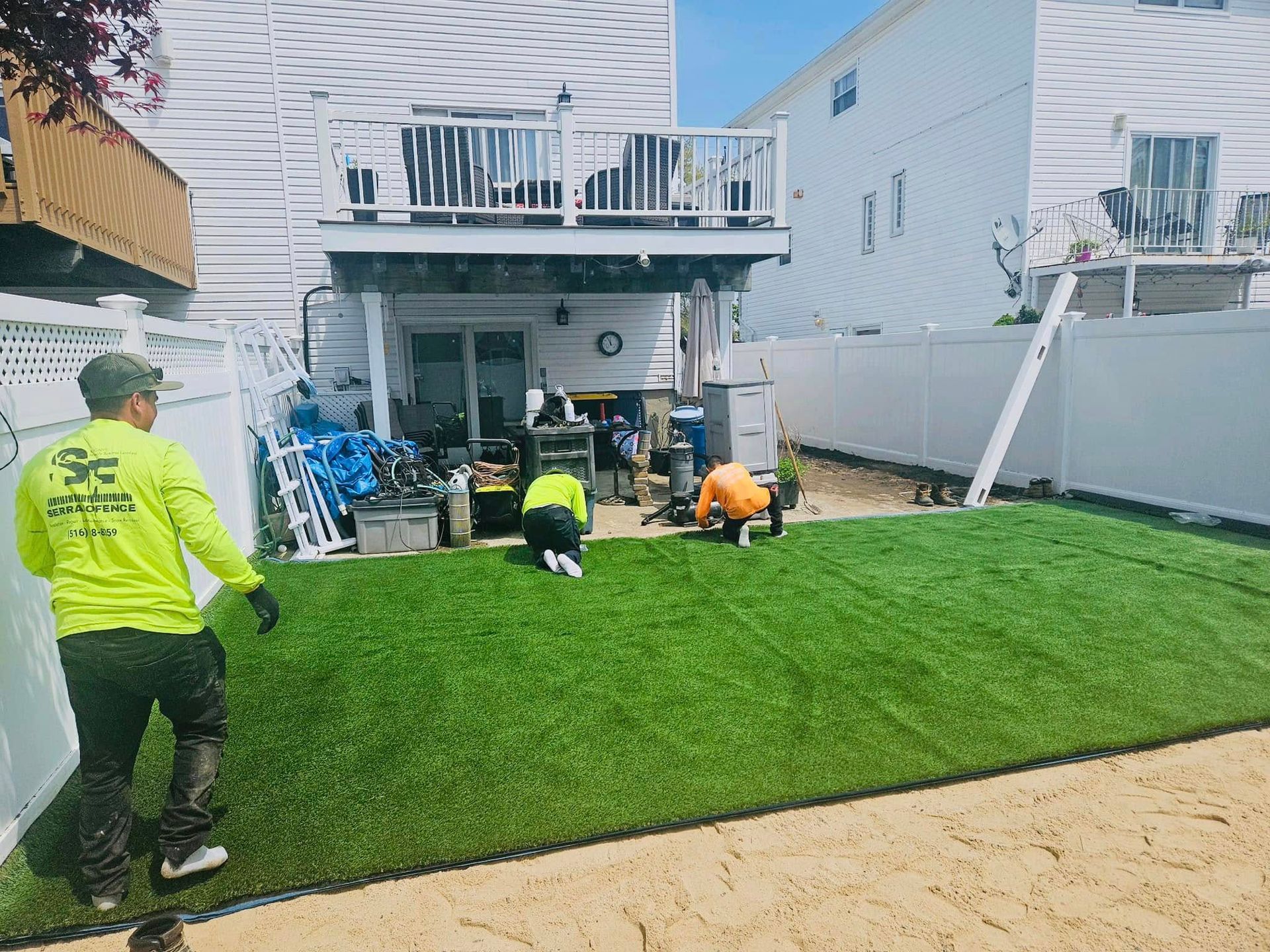 Workers install artificial turf in a backyard. Three people in safety vests on green grass, near a house and white fence.