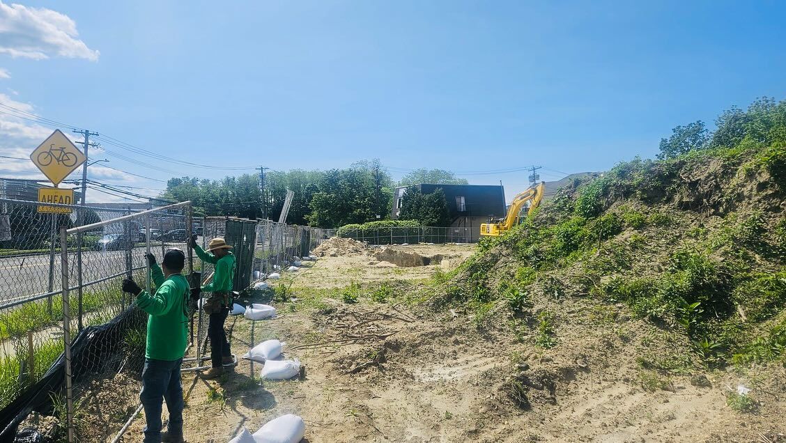 Two people in green shirts working on a chain-link fence on a sunny day. A sign and foliage are in the background.