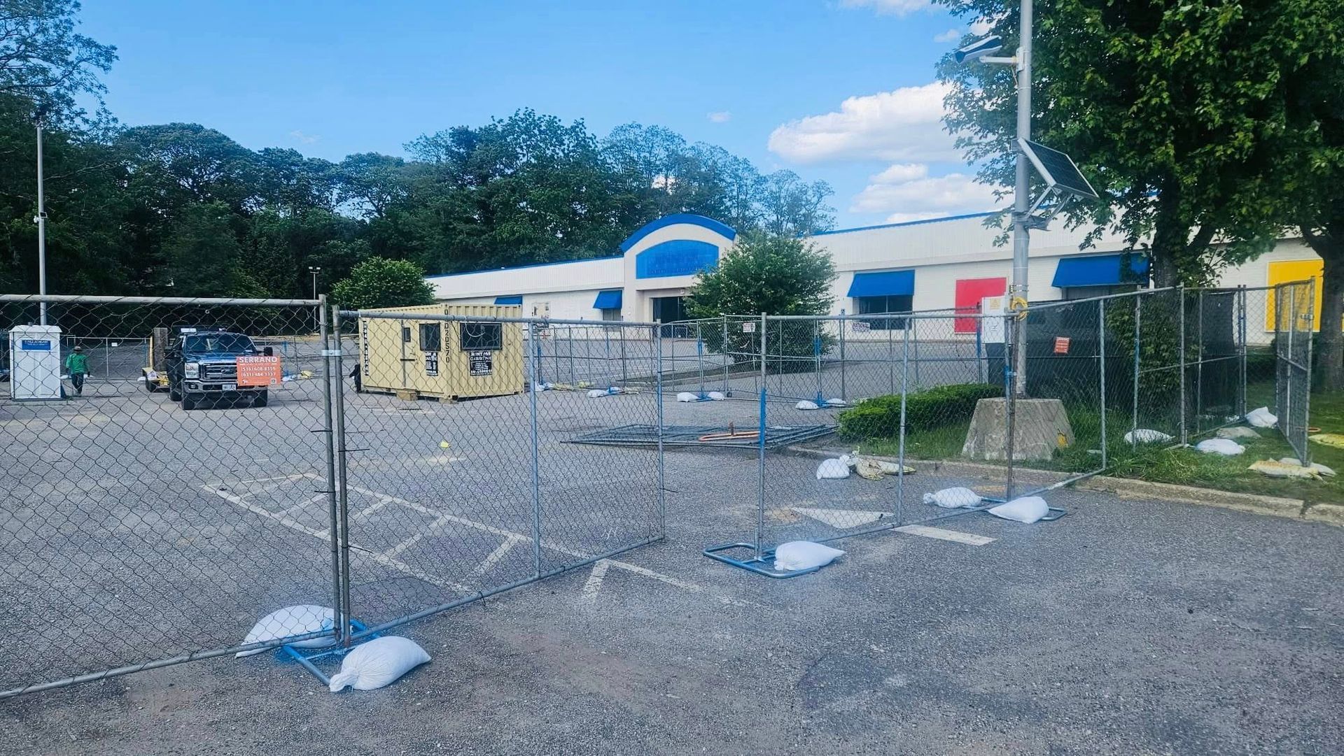 Construction site with a building, fenced in, and equipment. Blue and red awning is visible.