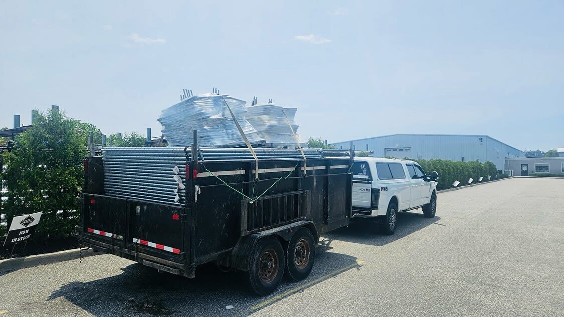 White truck towing a black trailer loaded with large, wrapped materials outside a building.
