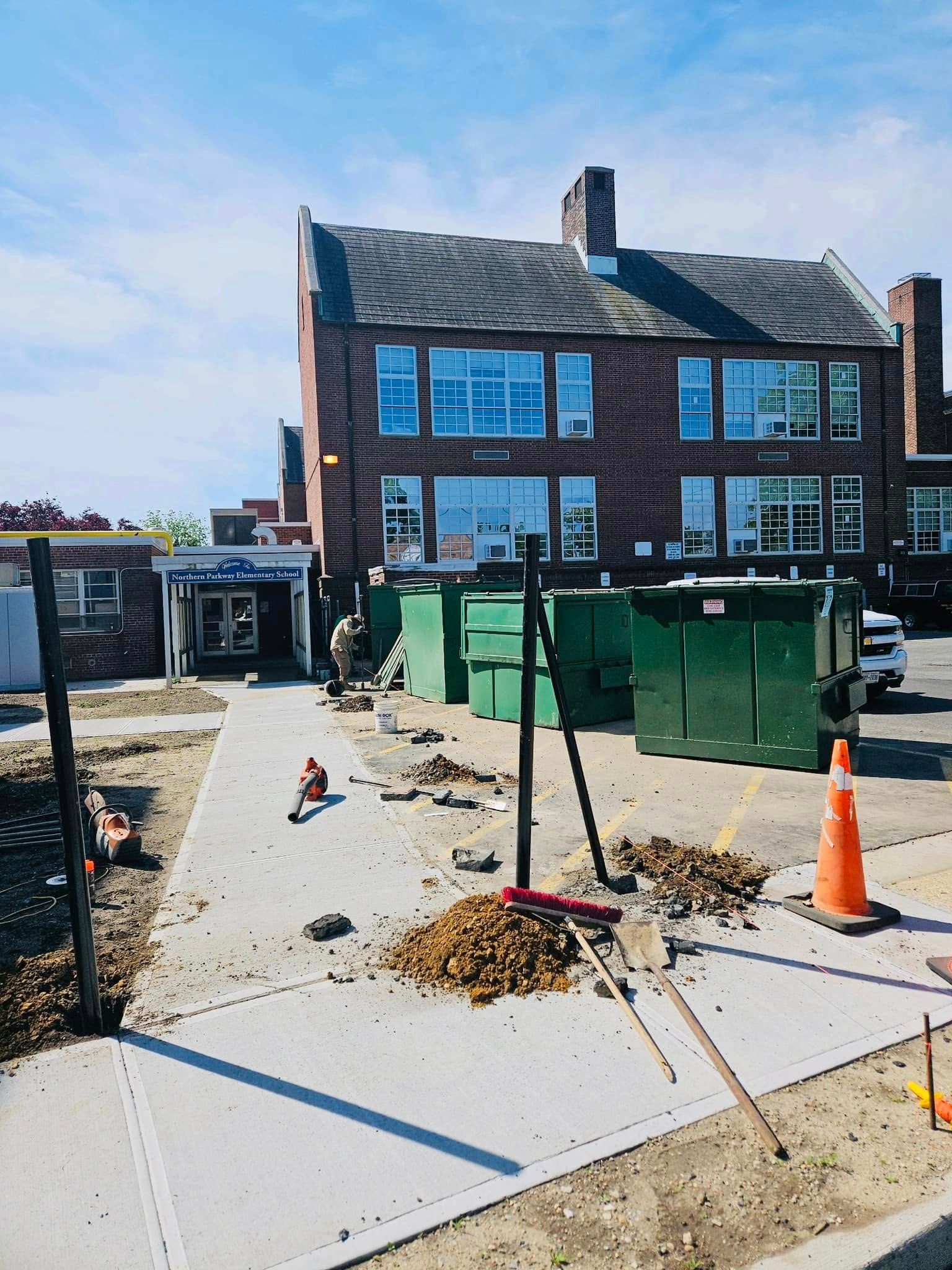 Construction site in front of a brick building. Green dumpsters, a sidewalk, and blue sky visible.