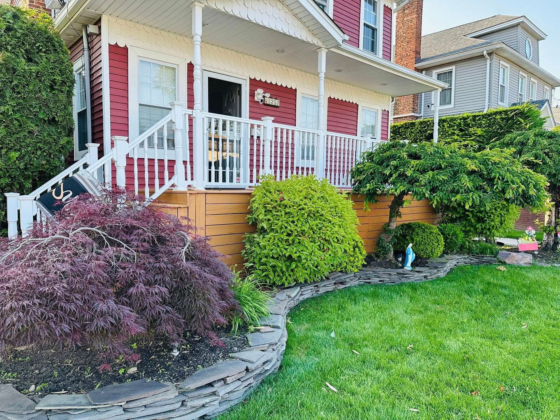 Red house with white porch, red shutters, and lush green lawn with a stone border.