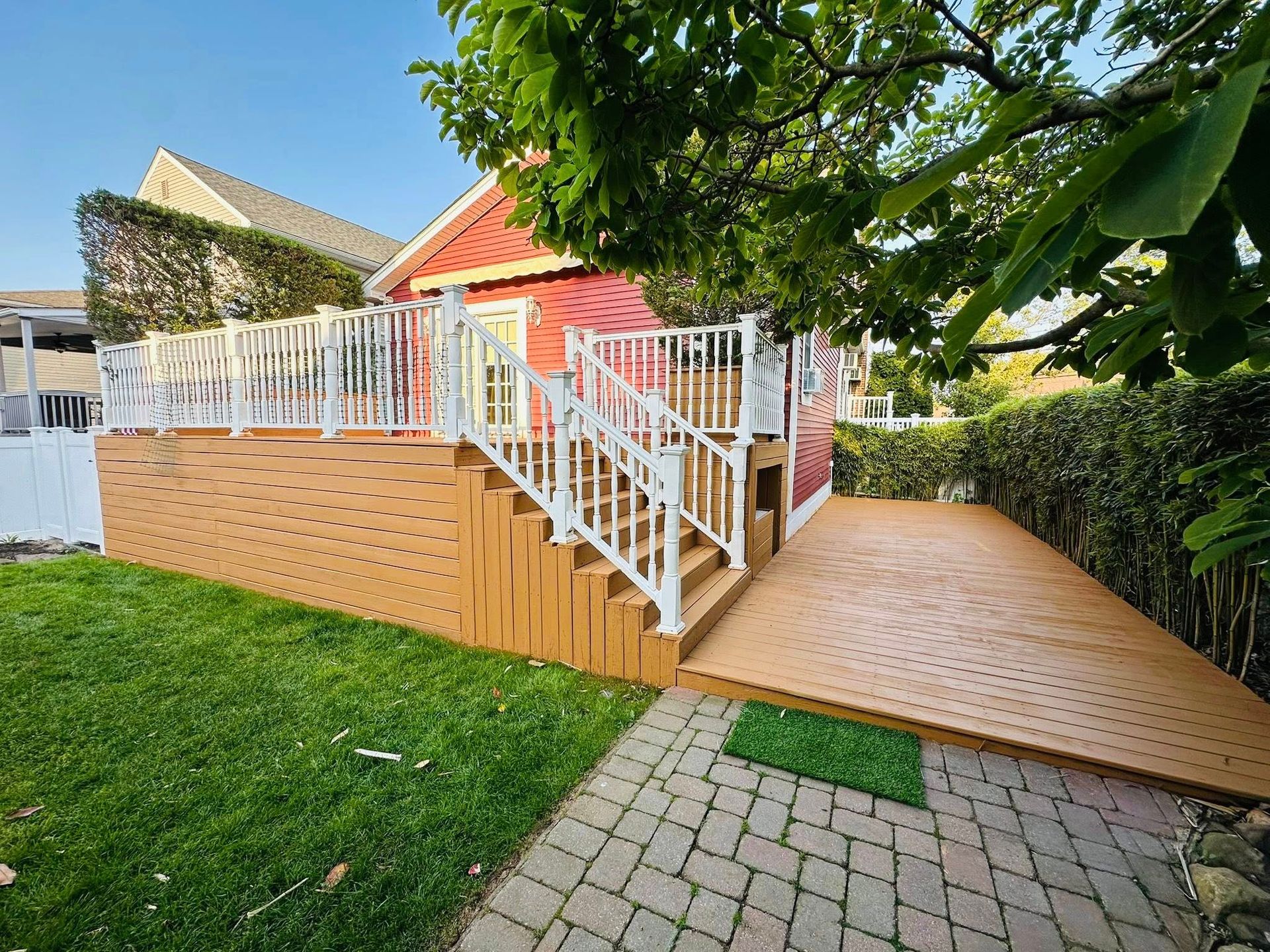 Red house with wooden deck, white railings, and stairs. Deck overlooks a paved patio on a green lawn.