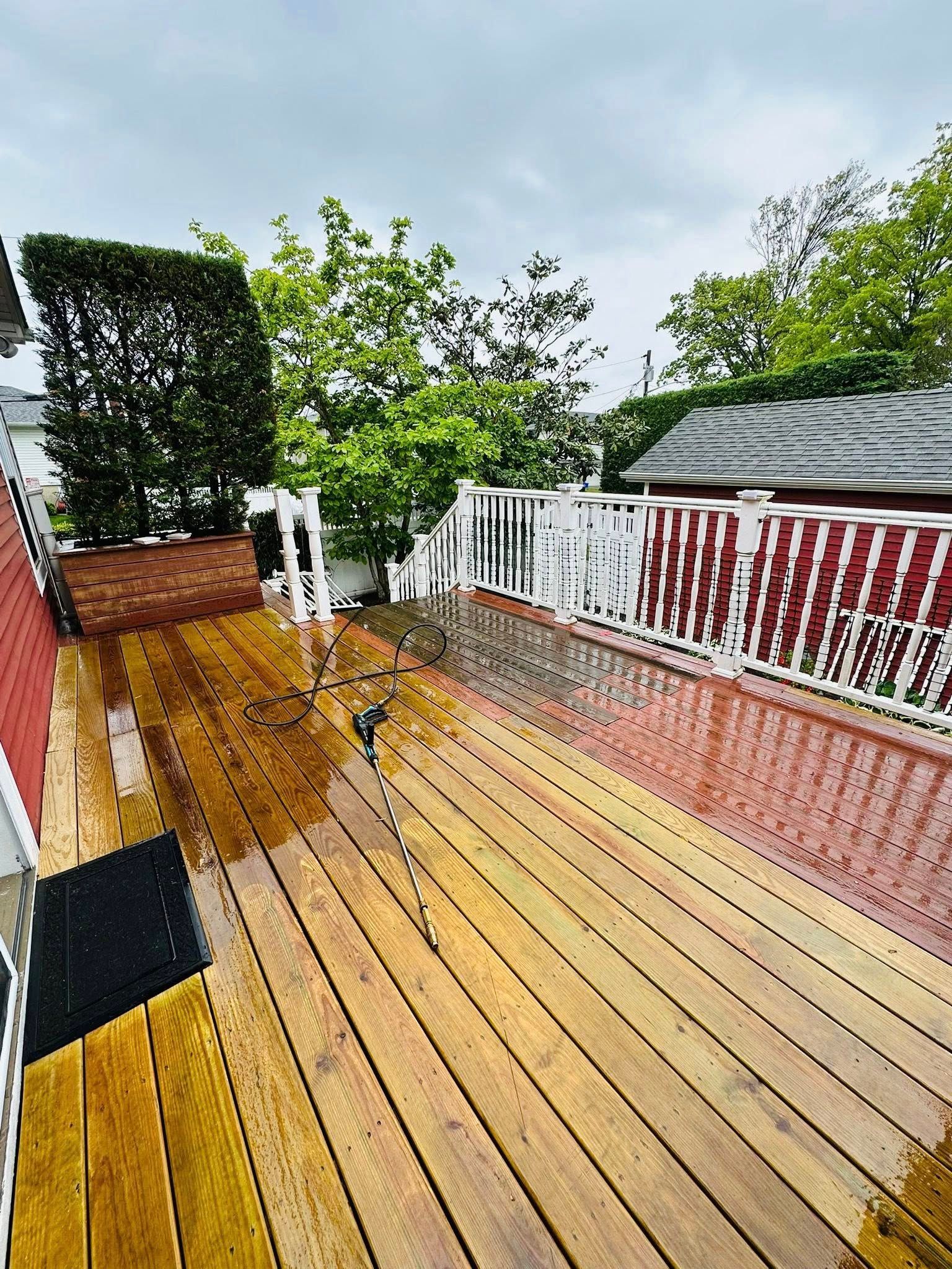 Wooden deck wet from rain, white railing, red building, green trees in background.