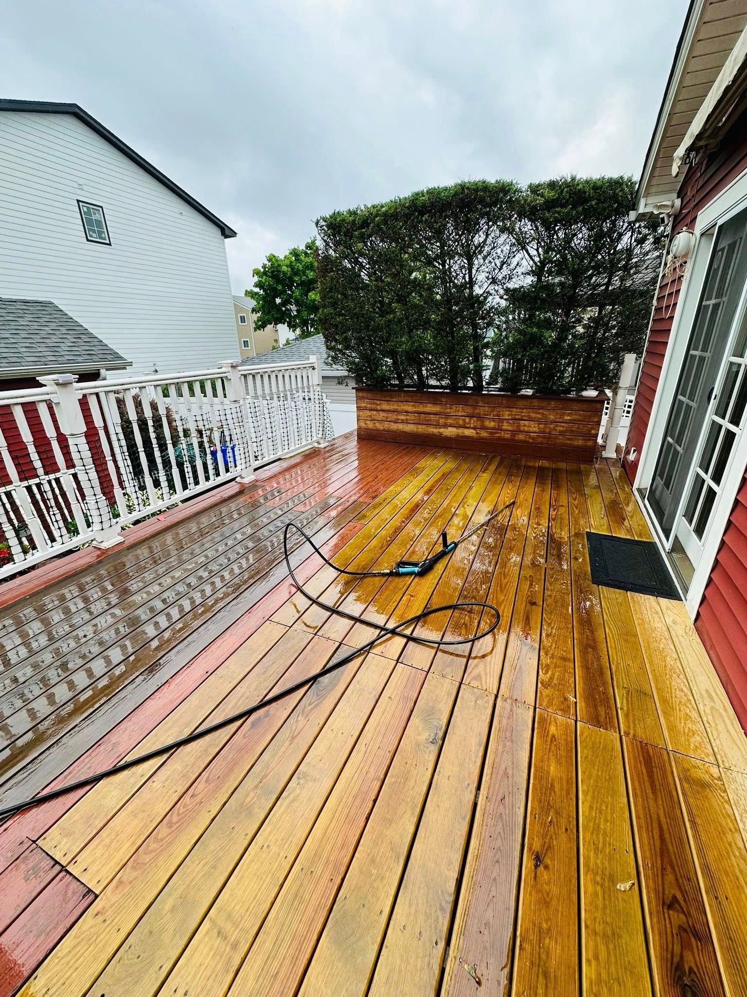 Wooden deck with wet boards, white railing, red house, and green hedge.