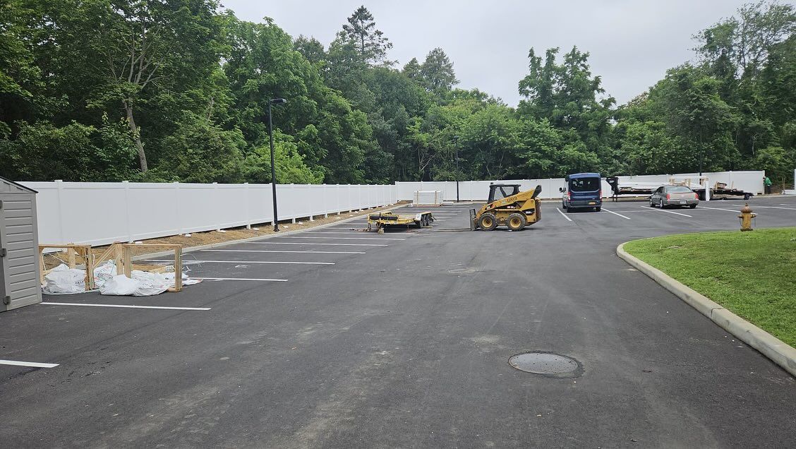 Construction site with asphalt and white fence, a yellow bulldozer, and trees in the background.