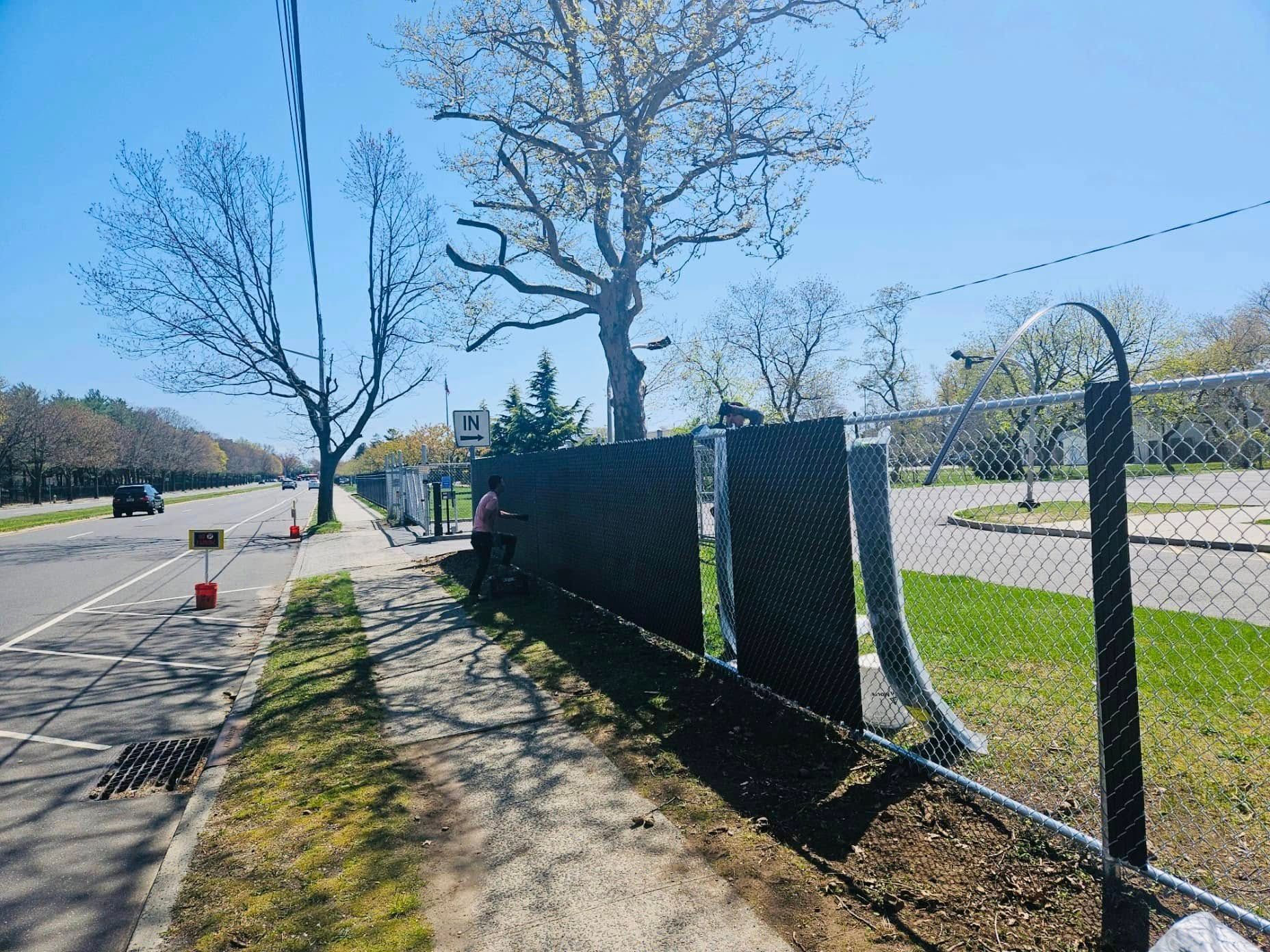 Fence with black privacy panels being installed along a road with a tree and blue sky.
