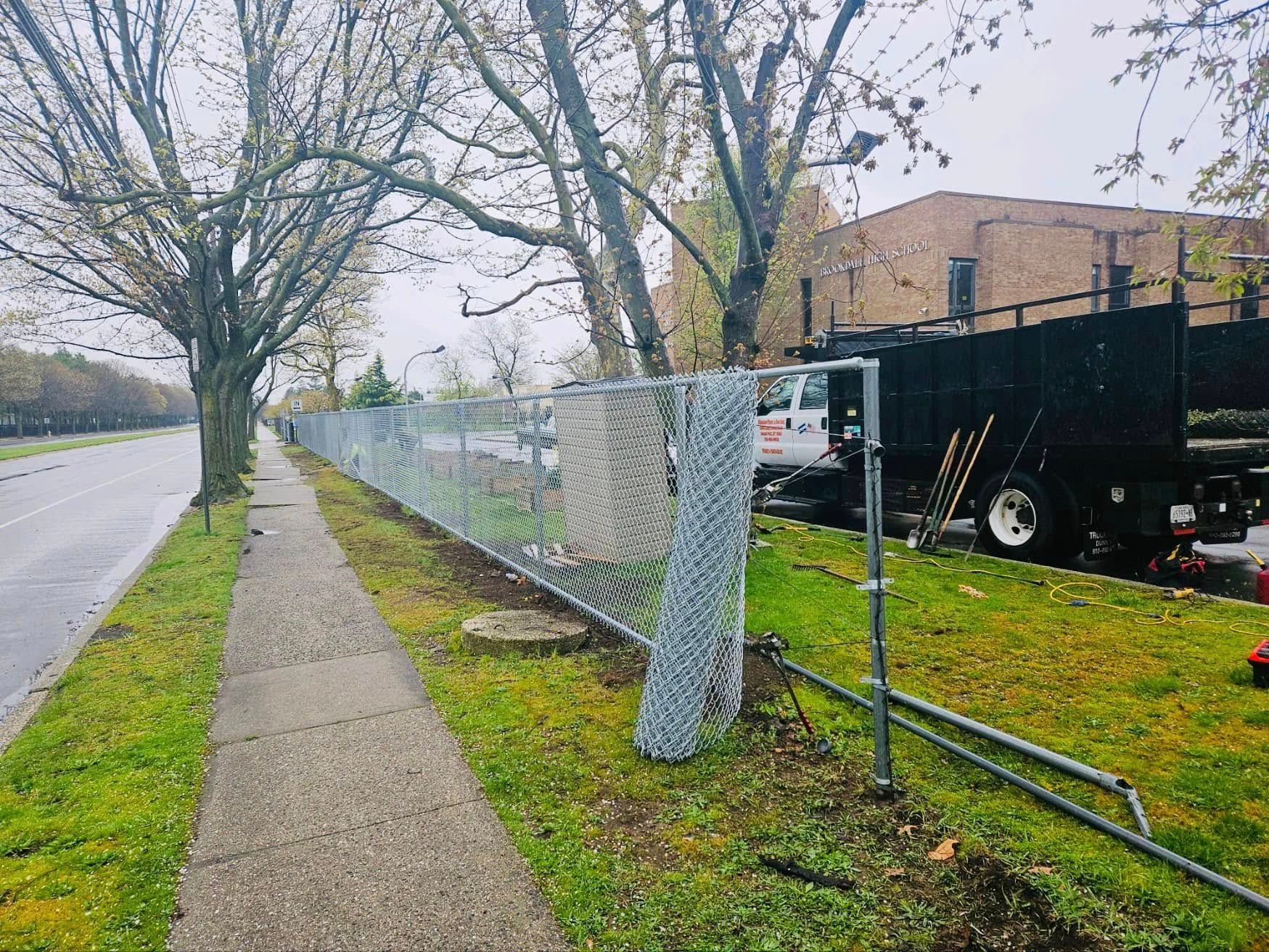 Chain-link fence damaged on a grassy area next to a sidewalk and road. A truck is parked nearby.