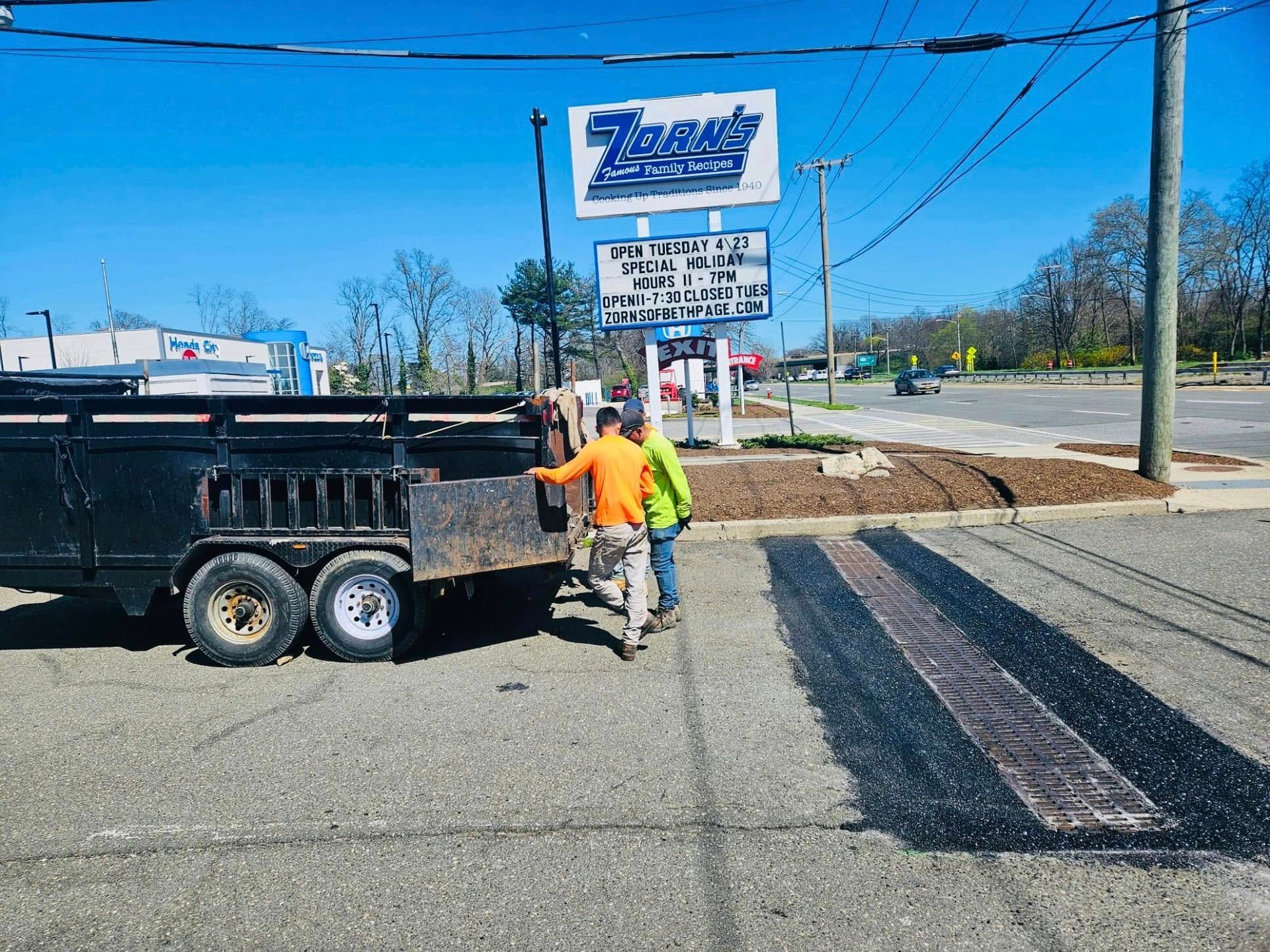 Two workers by a trailer next to a restaurant sign, near a road. Bright, sunny day.