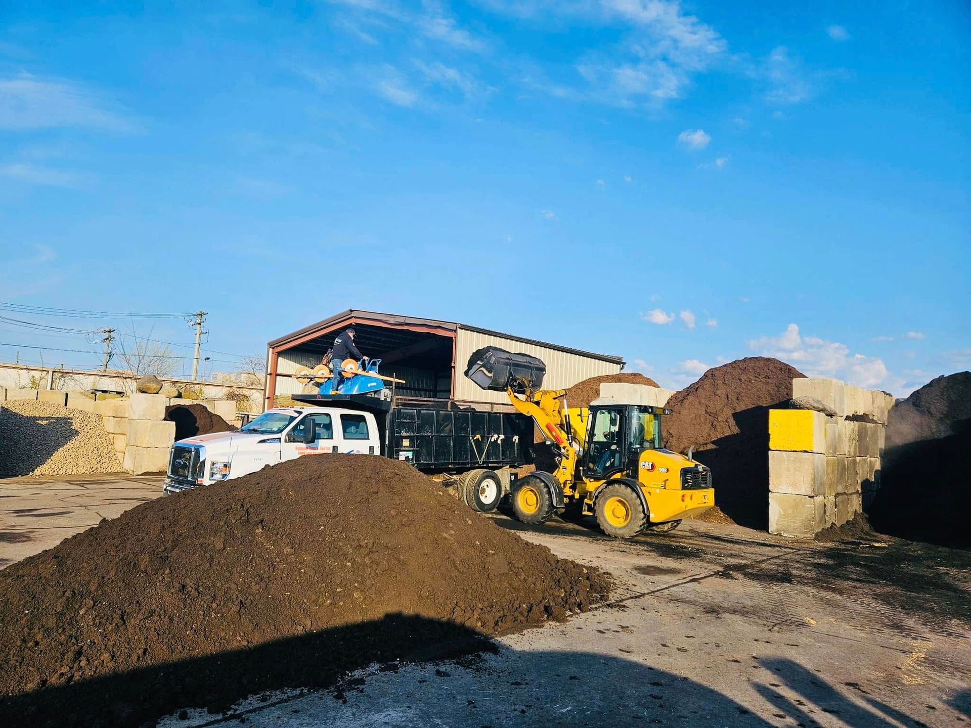 A yellow front-end loader loading a dump truck with compost at a facility under a blue sky.